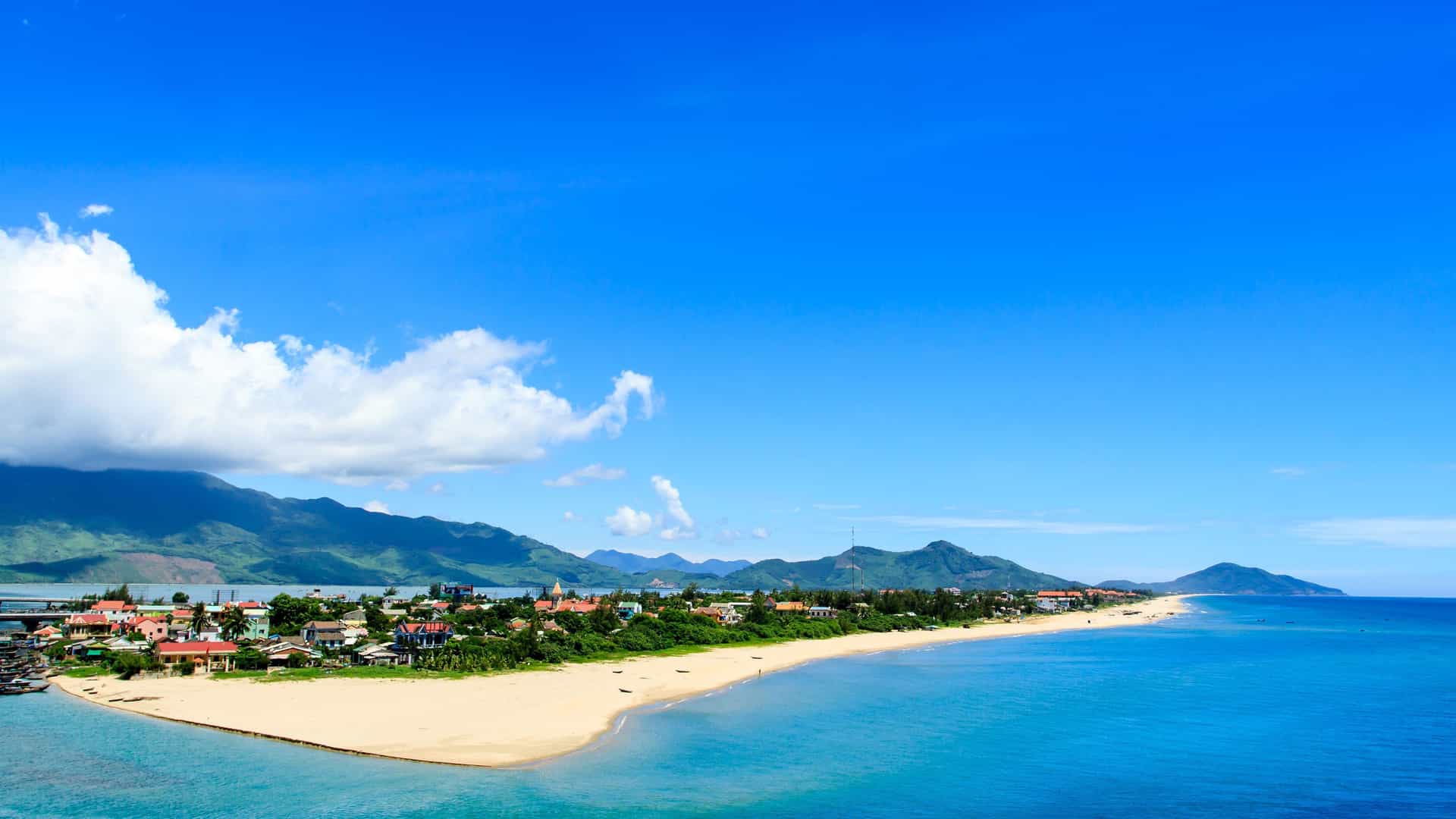 A panoramic shot of a beautiful beach and coastline in Hue, Vietnam, with a small town and mountains in the background, all under a vast, bright blue sky.