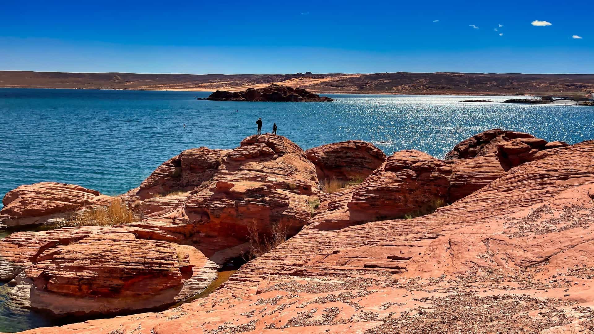 A scenic view of Sand Hollow State Park in Utah, featuring the stark contrast between the vibrant, warm blue waters of the Sand Hollow Reservoir and the surrounding red sandstone rock formations and crimson sand dunes of Sand Mountain. The image likely includes a person, or people, in the foreground or on a rock outcropping, looking out over the expansive, colorful landscape.