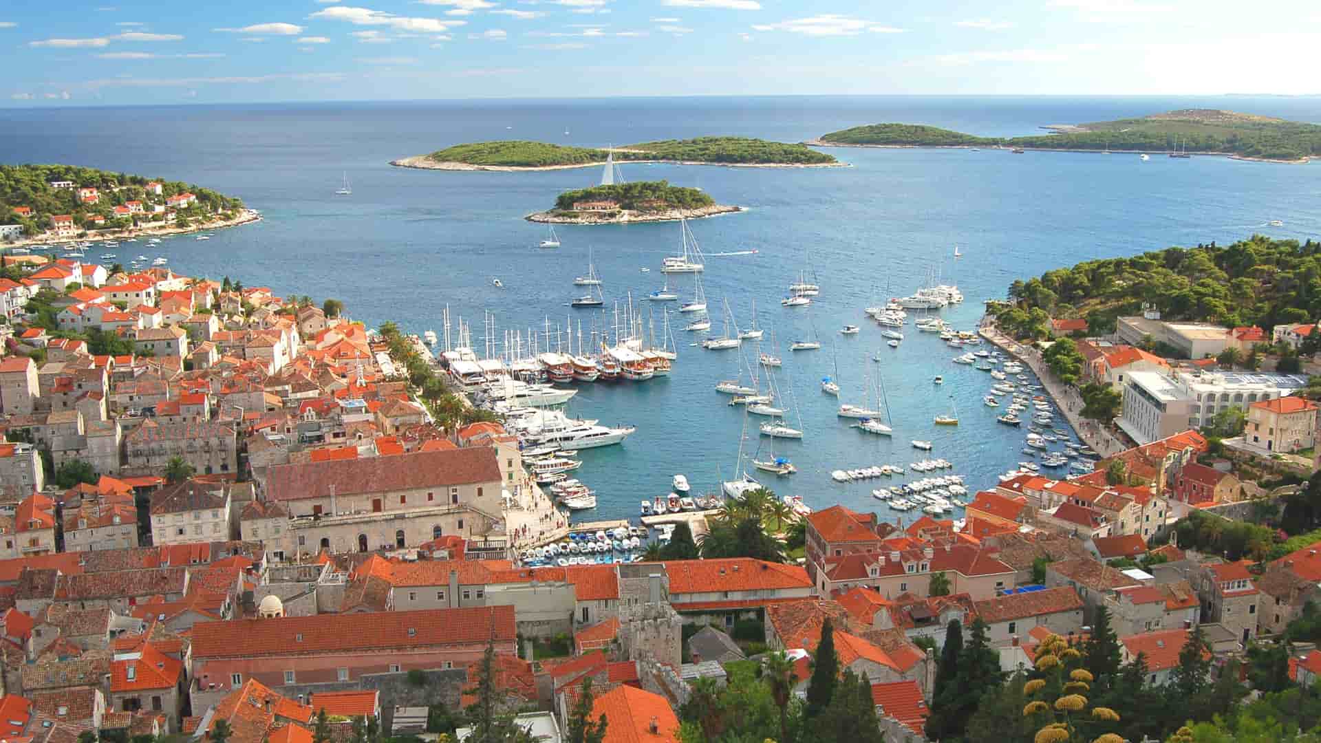 An aerial view of the picturesque harbor in Hvar, Croatia, with a bustling marina full of boats, red-roofed buildings of the town, and small, green islands dotting the turquoise sea.