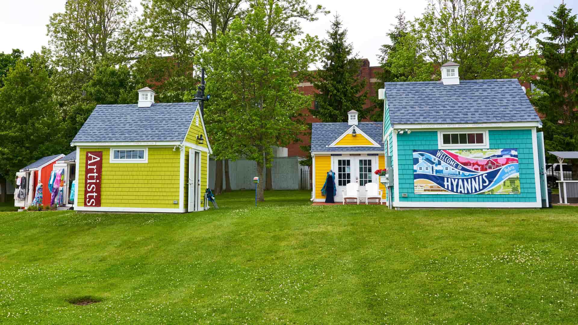 "A bright and cheerful row of colorful artist shacks on a green lawn with a large ""Welcome to Hyannis"" sign on one of the buildings in Hyannis, Massachusetts. "