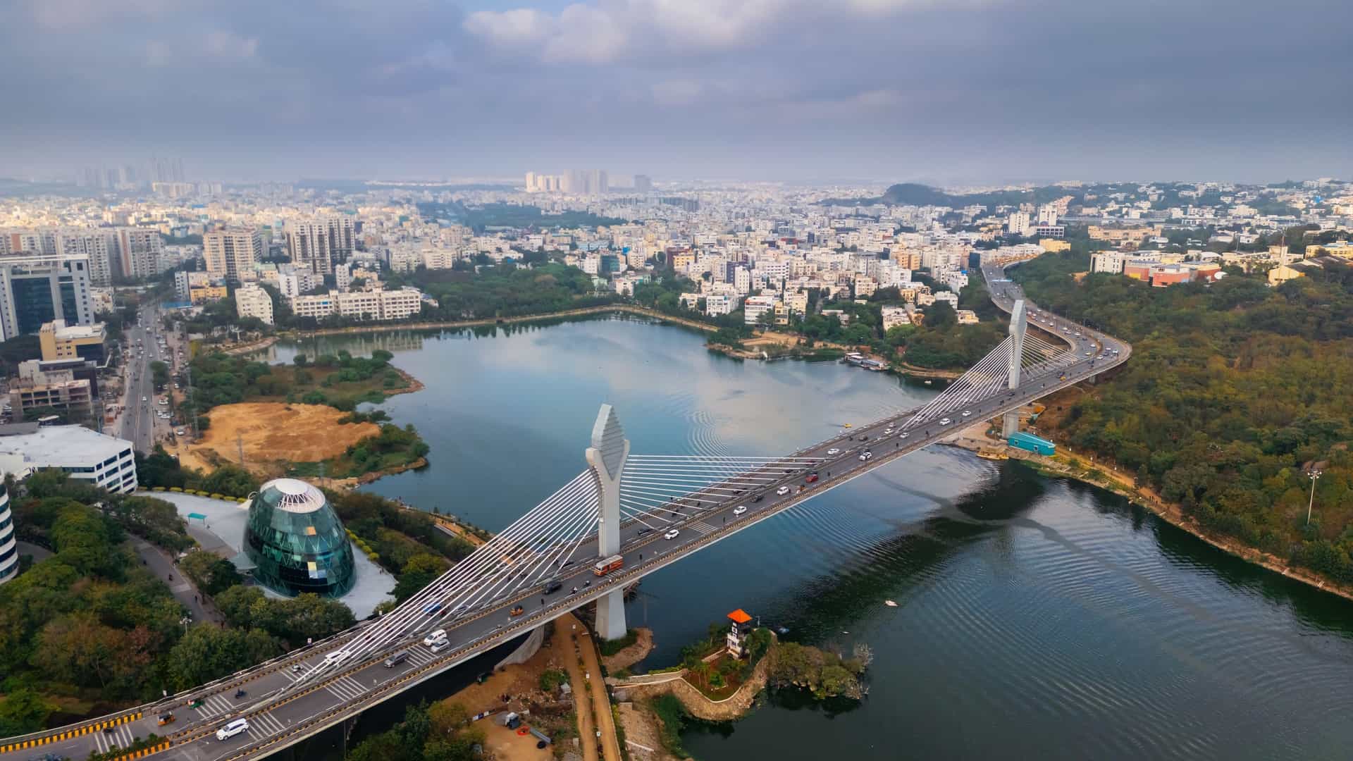 An aerial view of Hyderabad, India, showing the modern, cable-stayed Durgam Cheruvu Bridge crossing the Durgam Cheruvu lake, with a sprawling cityscape and commercial buildings in the distance.