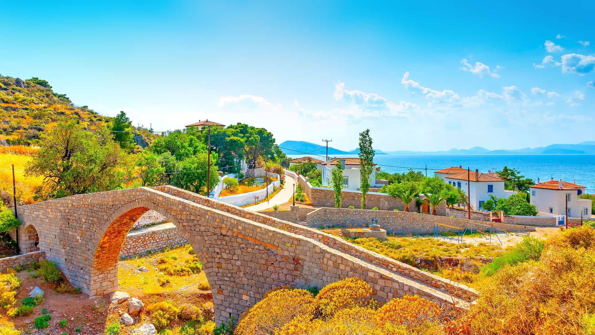 A picturesque stone bridge on the Greek island of Hydra, with a winding road leading to whitewashed houses and the sparkling Aegean Sea under a clear blue sky.