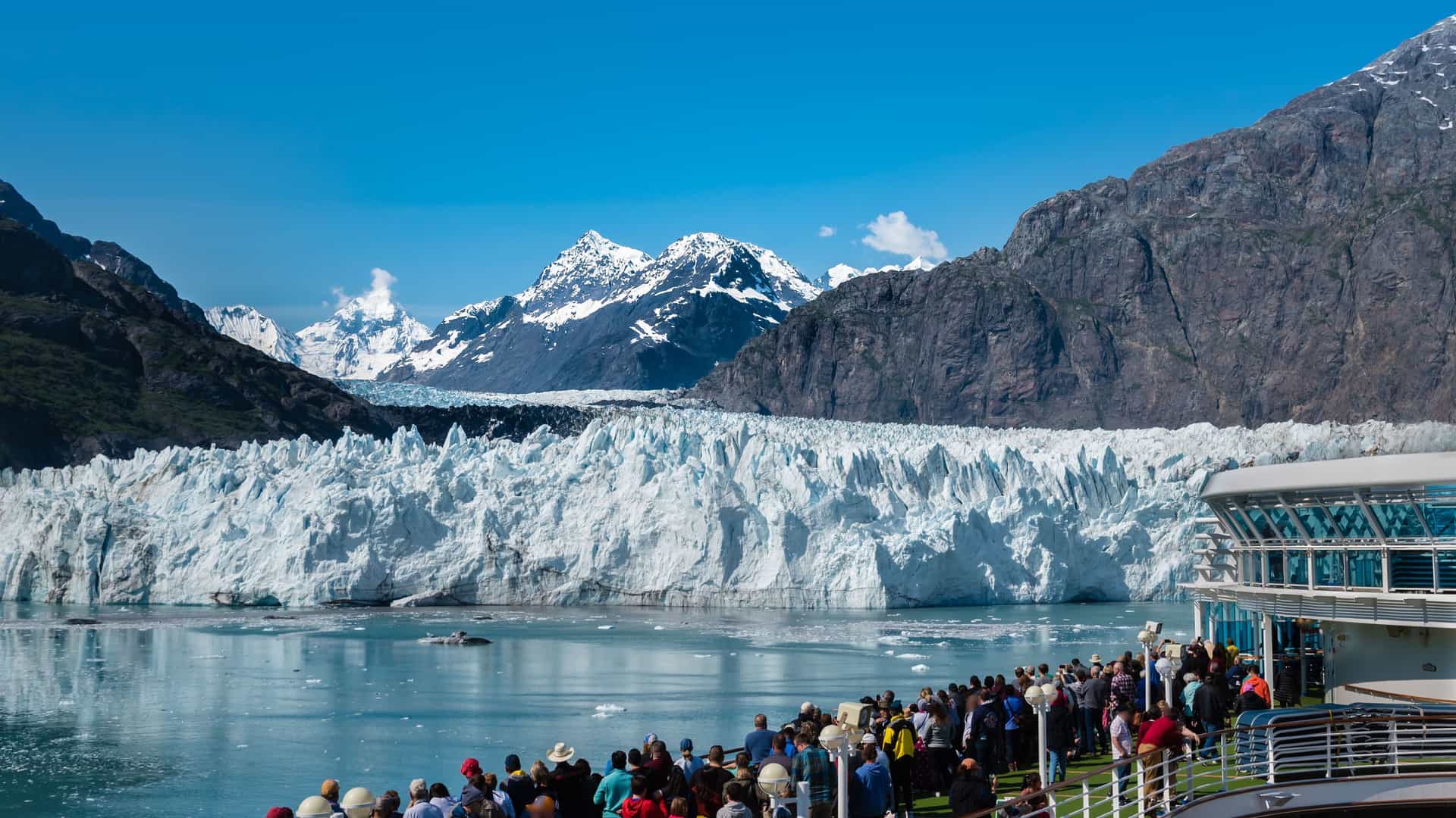 A scenic view from the deck of a cruise ship, with passengers admiring a massive, jagged glacier and snow-capped mountains during an ice edge cruising experience.