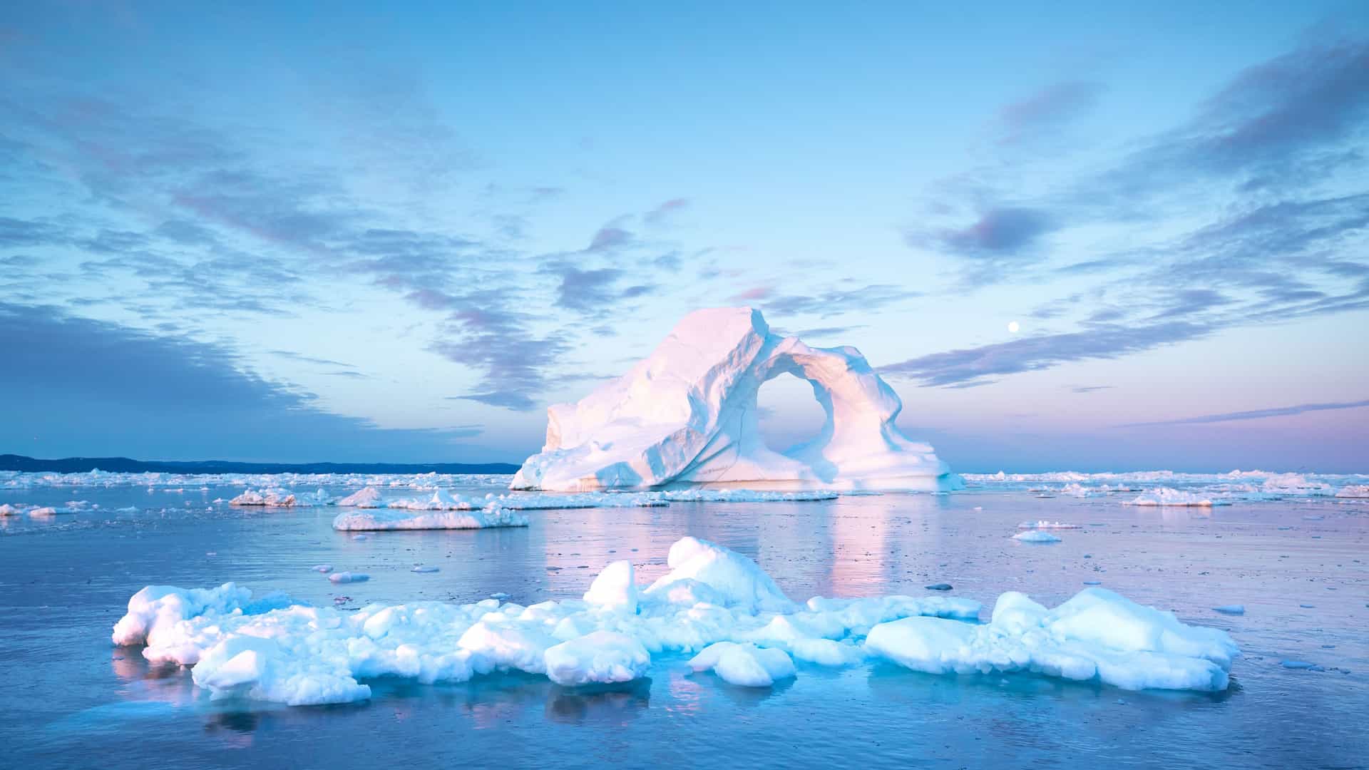 A serene polar landscape with a large, arch-shaped iceberg in Icy Bay, Alaska, surrounded by smaller ice floes floating in the calm water under a pink and blue sky.