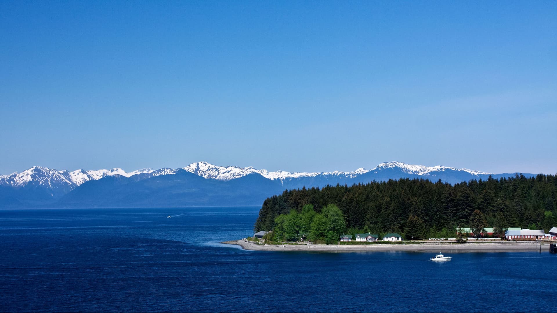 A serene coastal landscape of Icy Strait Point, Alaska, with a small settlement nestled among a dense pine forest, a calm blue sea, and a stunning backdrop of snow-capped mountains.