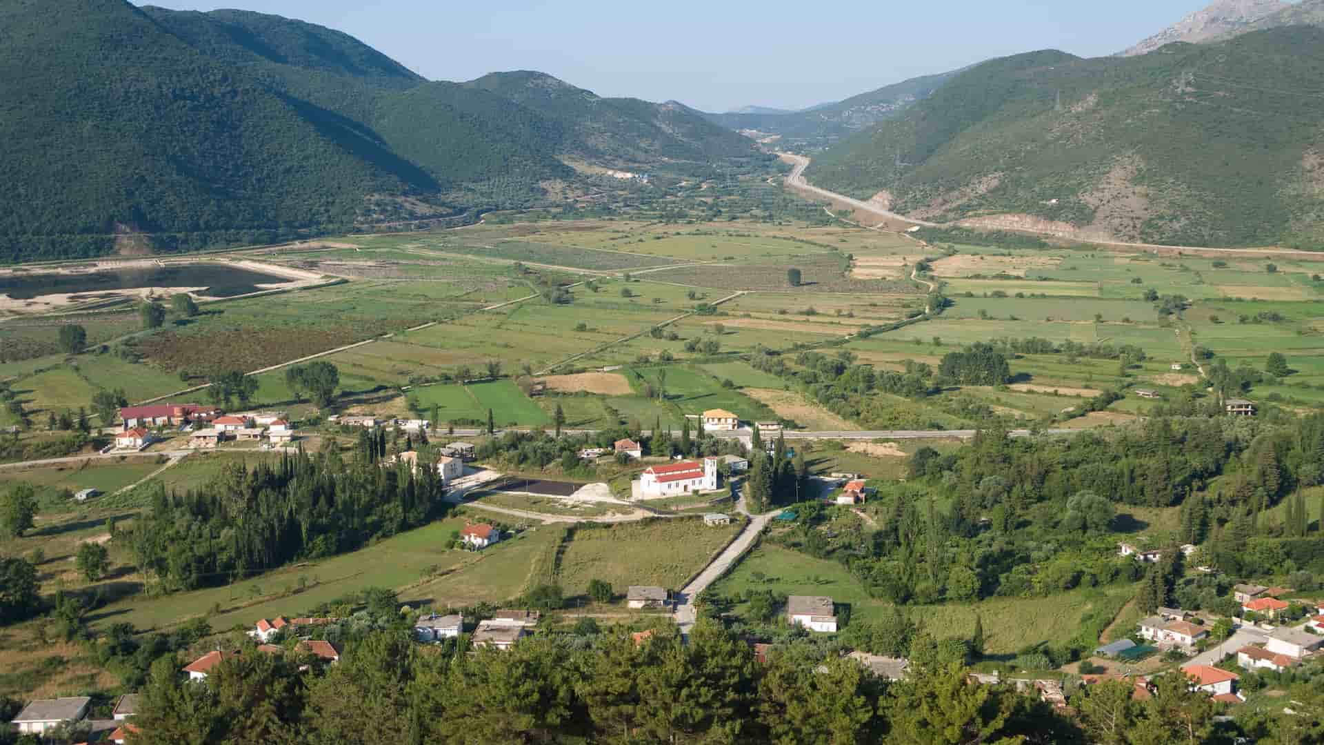 A scenic aerial view of the lush, green valley and small village in Igoumenitsa, Greece, with mountains in the background and fields of farmland stretching to the horizon.