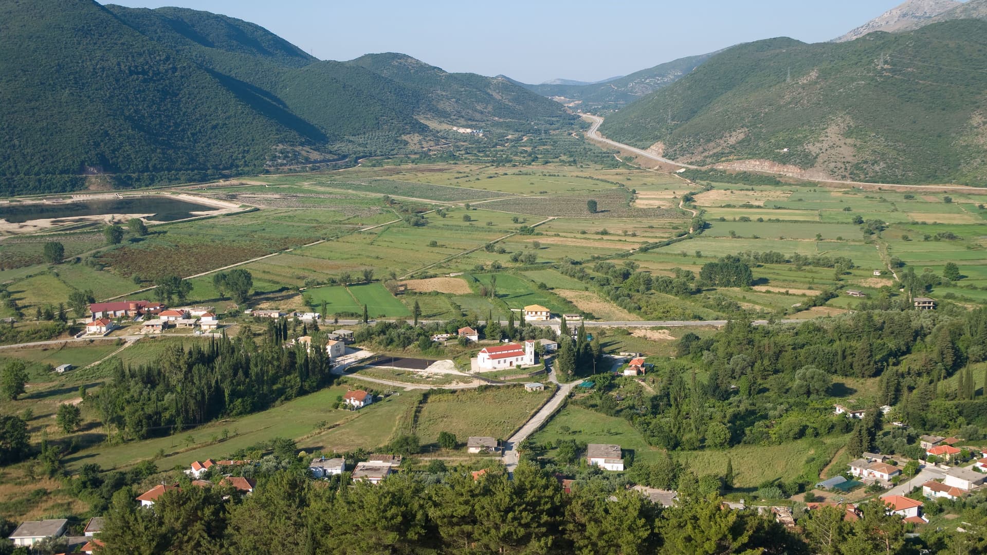 A scenic aerial view of the lush, green valley and small village in Igoumenitsa, Greece, with mountains in the background and fields of farmland stretching to the horizon.
