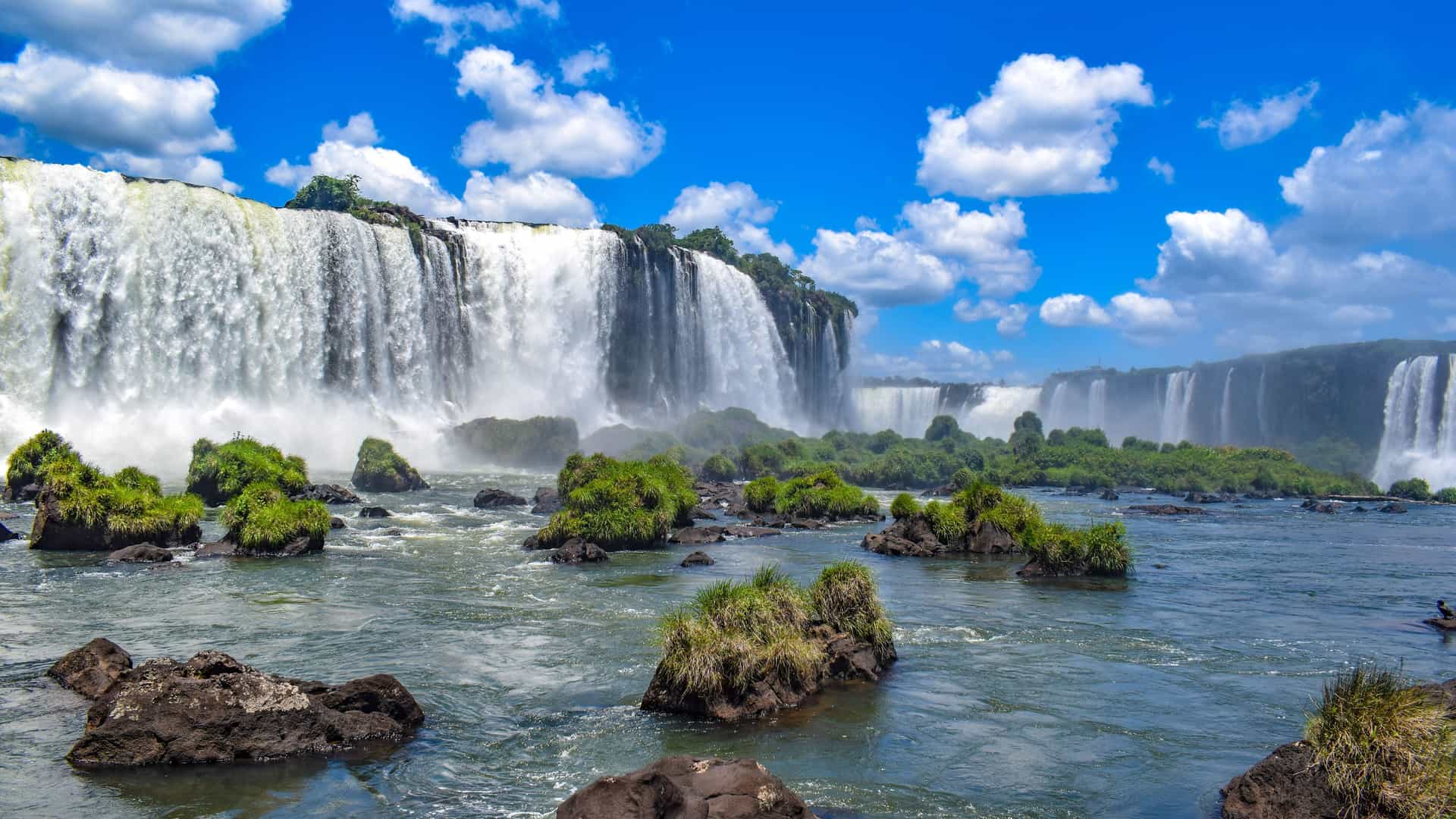 A powerful view of the majestic Iguassu Falls on the Brazilian side, with massive cascades of water, lush green foliage on the rocks, and a vibrant blue sky.