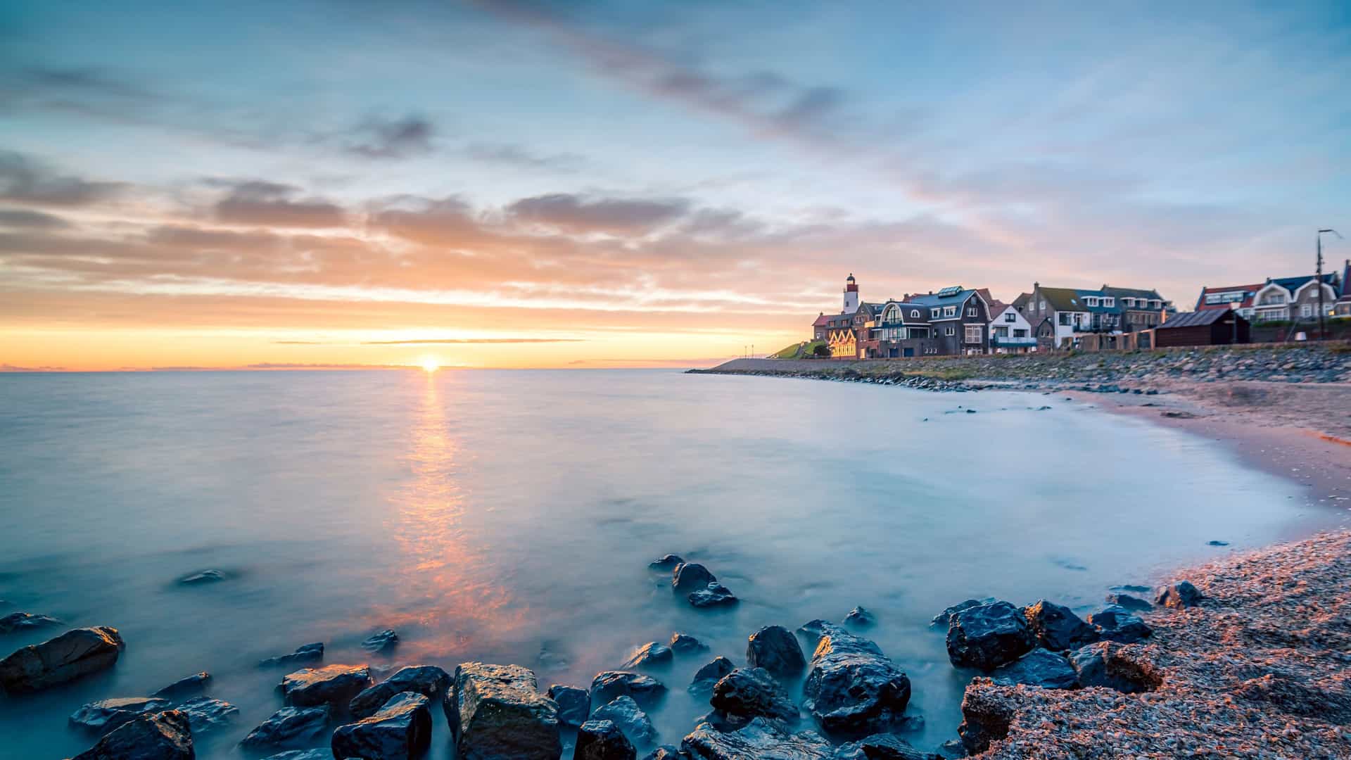 A breathtaking sunset over the calm waters of Ijsselmeer in the Netherlands, with the sun setting behind a small fishing village and a rocky beach in the foreground.