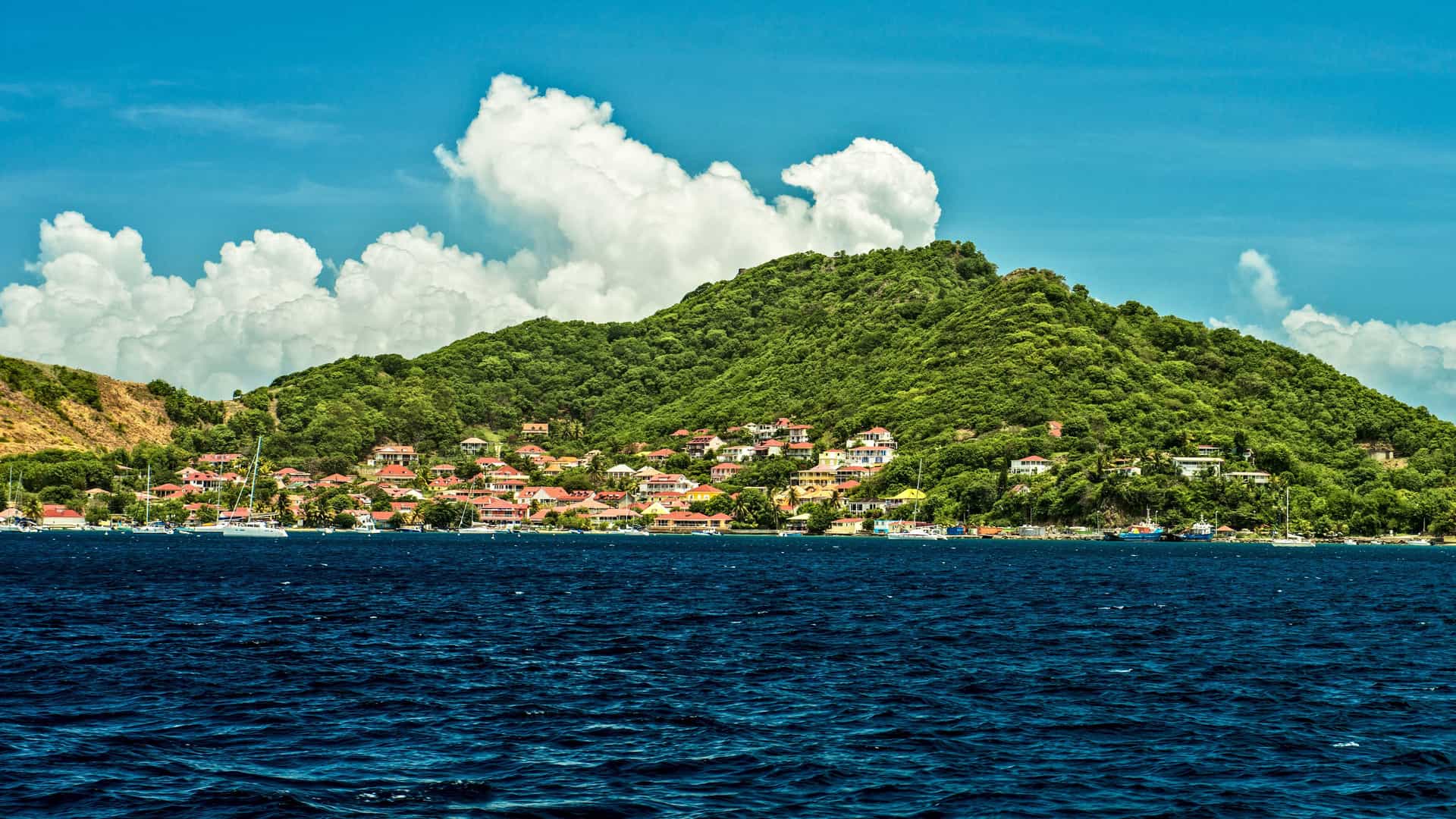 A picturesque view of a small town nestled at the base of a lush, green mountain on Iles des Saintes, Guadeloupe, with boats moored in the deep blue water and fluffy clouds in the sky.