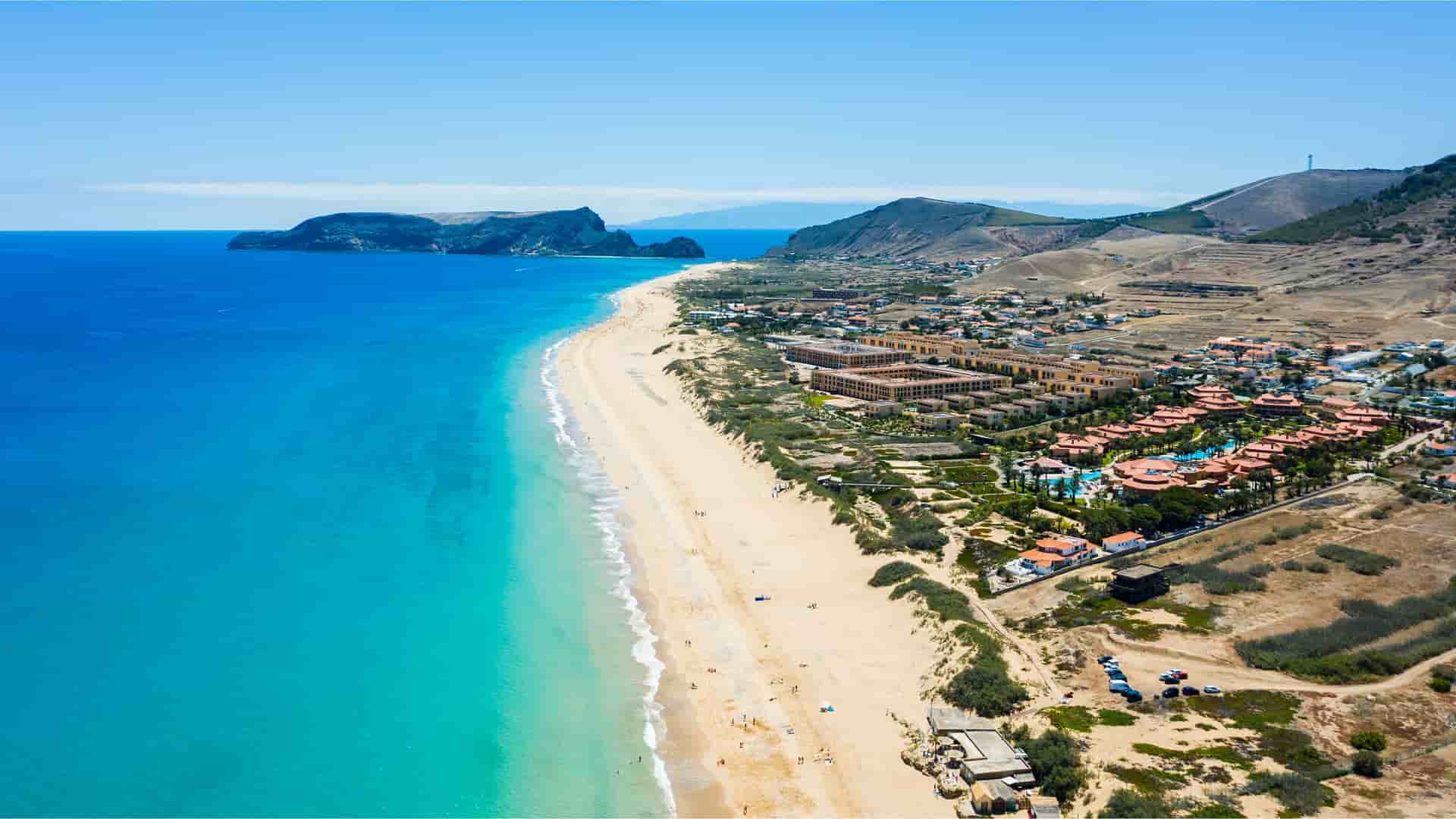 "An aerial view of Porto Santo Island, Portugal, showing the long, sandy beach and the turquoise waters of the Atlantic Ocean, with resorts and buildings nestled at the base of the island's mountains. "