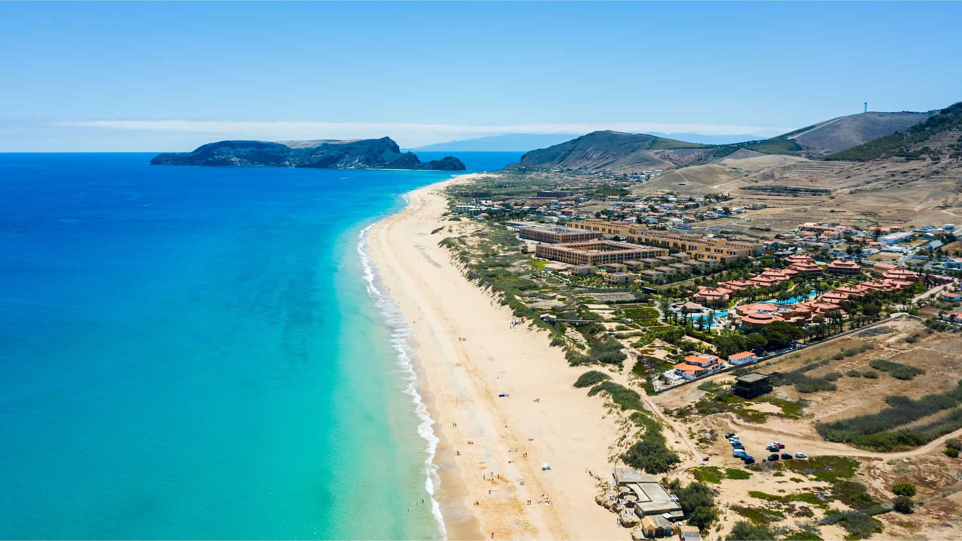 "An aerial view of Porto Santo Island, Portugal, showing the long, sandy beach and the turquoise waters of the Atlantic Ocean, with resorts and buildings nestled at the base of the island's mountains. "