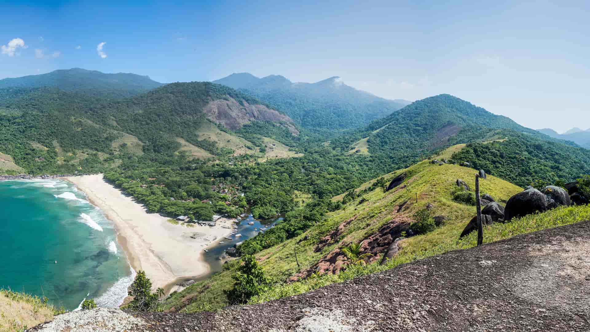 A scenic panoramic view of Ilhabela, Brazil, showing a long, sandy beach with turquoise water, a river flowing to the sea, and lush, green mountains under a bright blue sky.