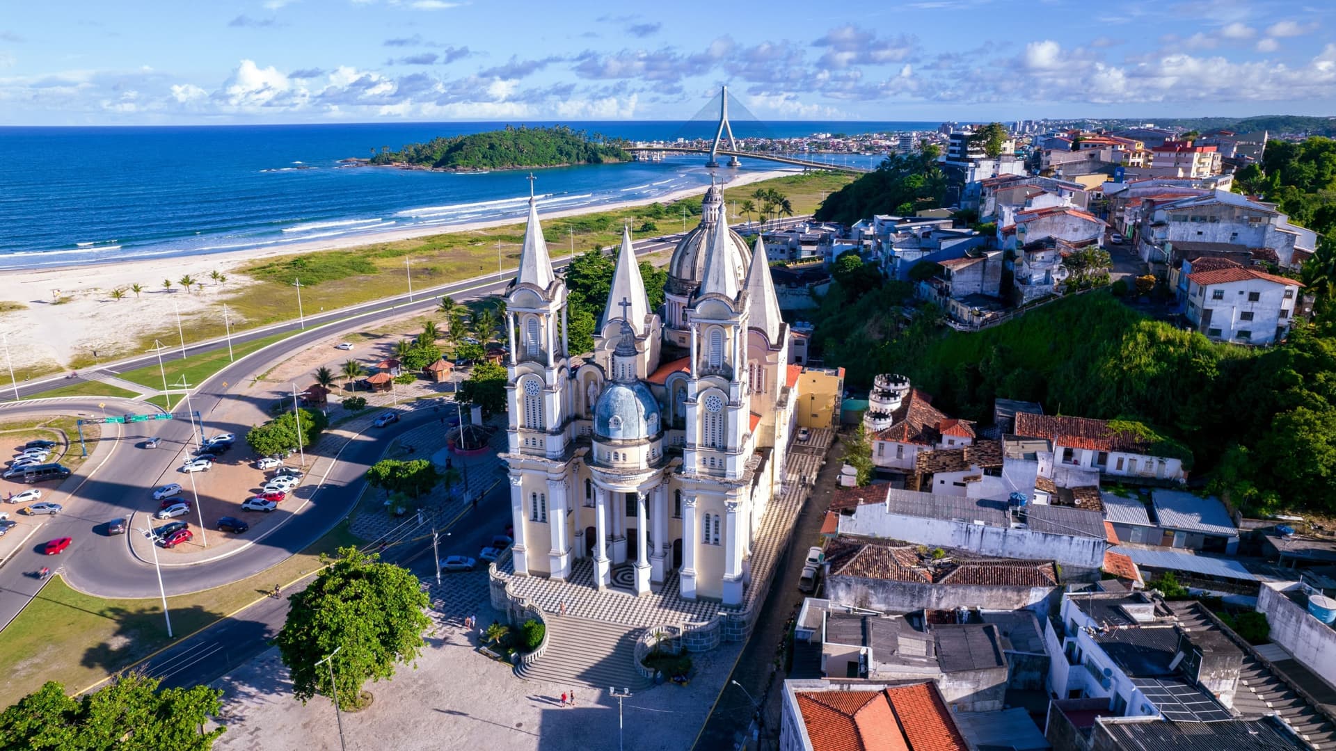 An aerial or high-angle view of Ilhéus, a coastal city in the state of Bahia, Brazil. The historic city center is visible, featuring a dense grid of buildings, with the prominent white structure of the São Sebastião Cathedral and the confluence of a river and the Atlantic Ocean. The image captures the contrast between the urban development and the surrounding tropical landscape.