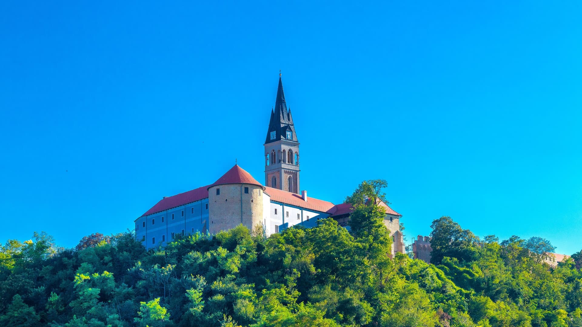 A majestic shot of Ilok Castle in Croatia, showcasing the large fortress with its prominent bell tower and red-roofed buildings perched on a lush, green hill under a clear blue sky.