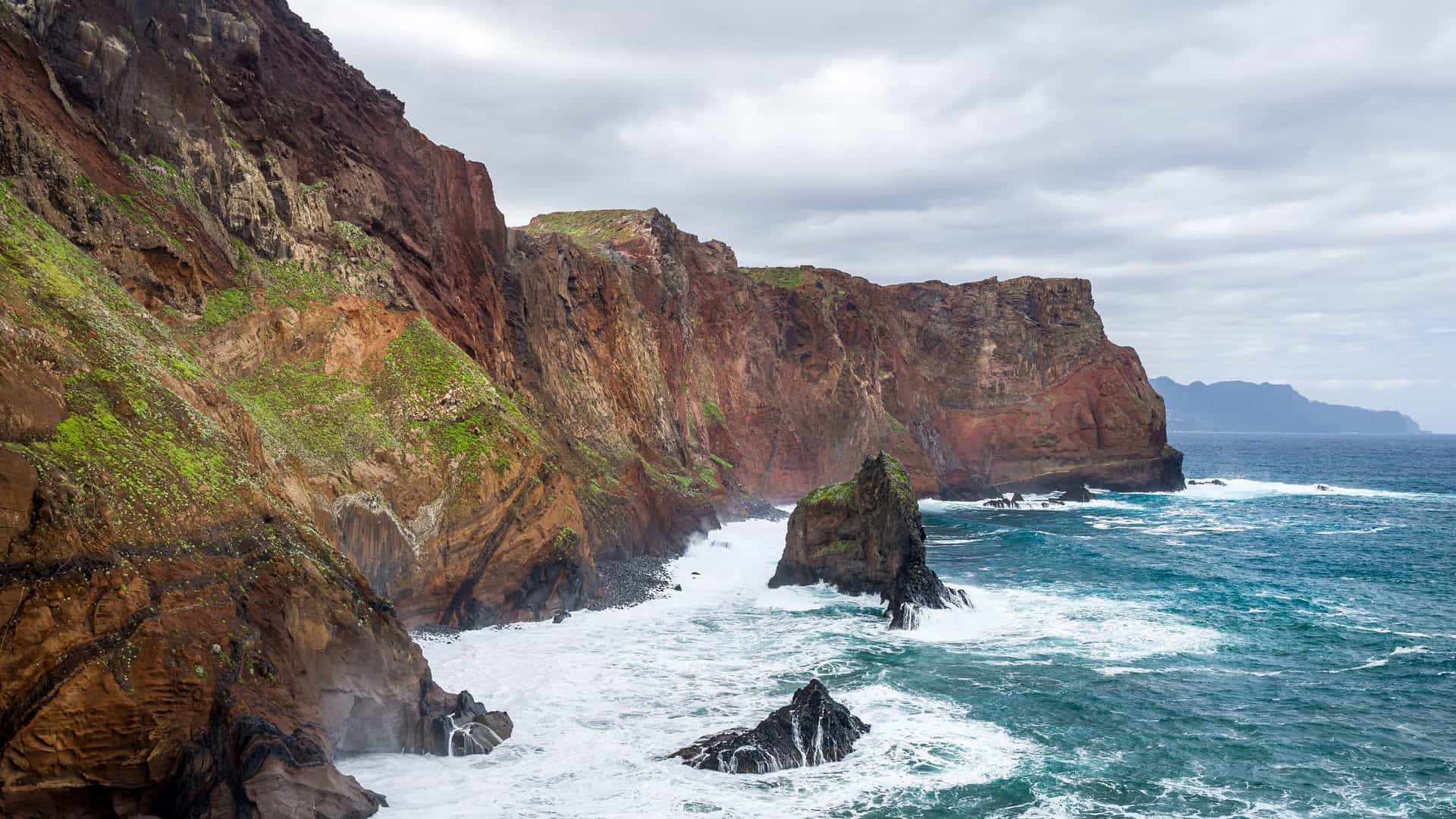 A stunning view of the steep, rocky cliffs and dramatic coastline of Inaccessible Island, with powerful waves crashing against the shore and a gray, cloudy sky.