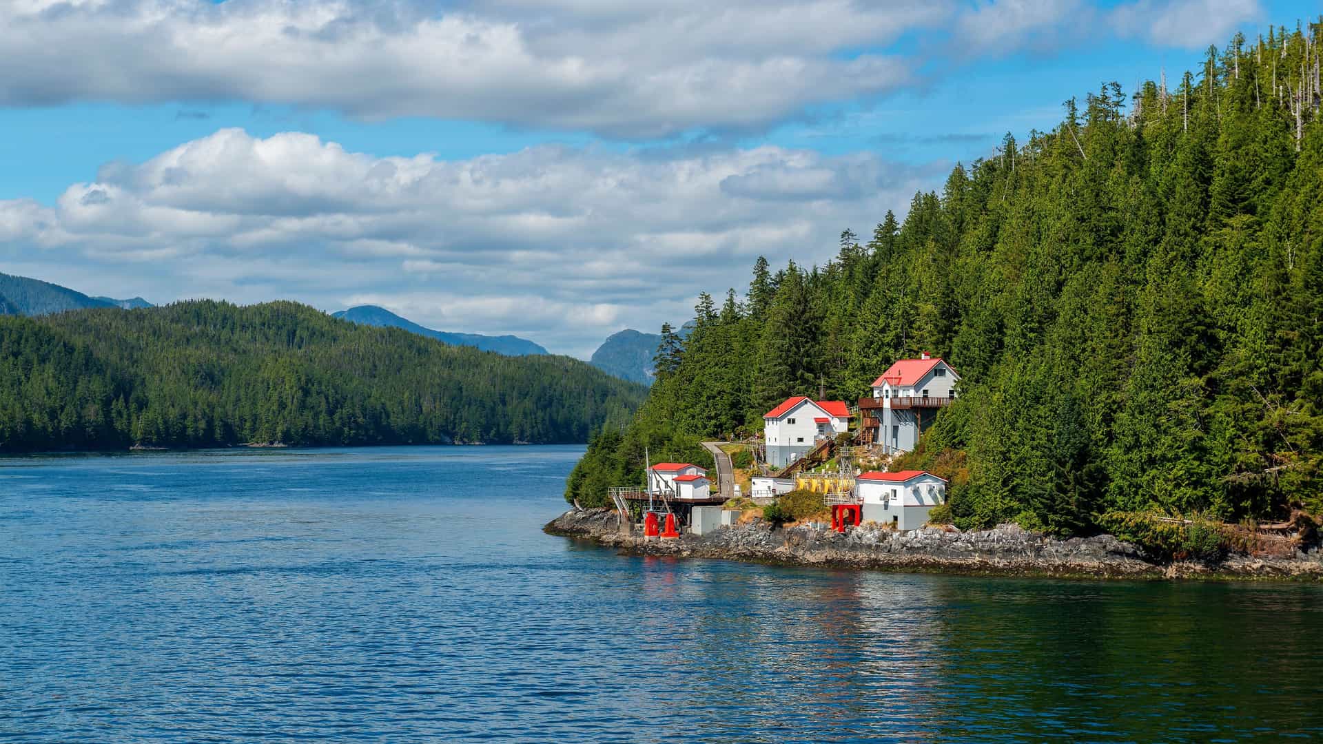 A serene view of the Inside Passage in Alaska, featuring a small group of white and red-roofed buildings on a rocky shoreline, surrounded by a dense green pine forest and calm blue water.