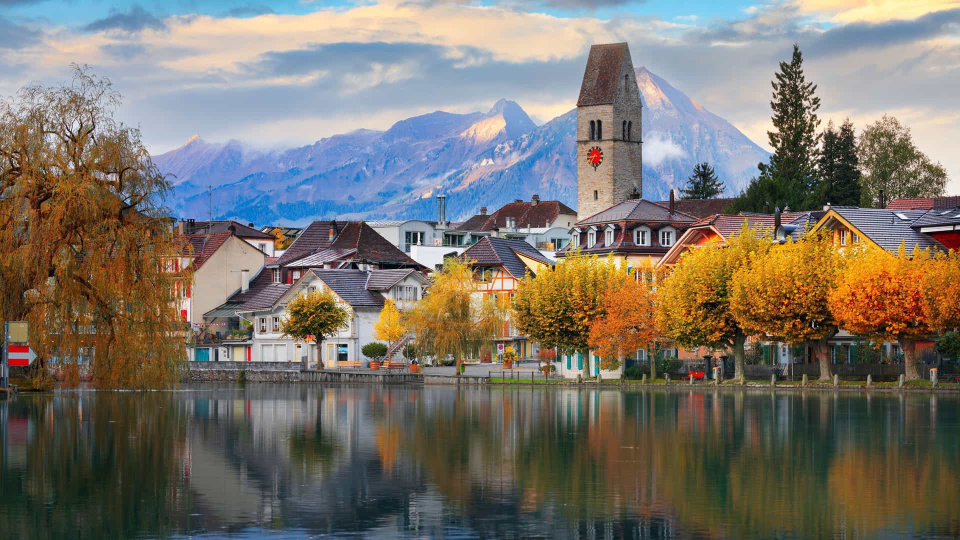 A picturesque view of Interlaken, Switzerland, with a peaceful lake reflecting the colorful buildings and autumn trees, all framed by majestic snow-capped mountains.