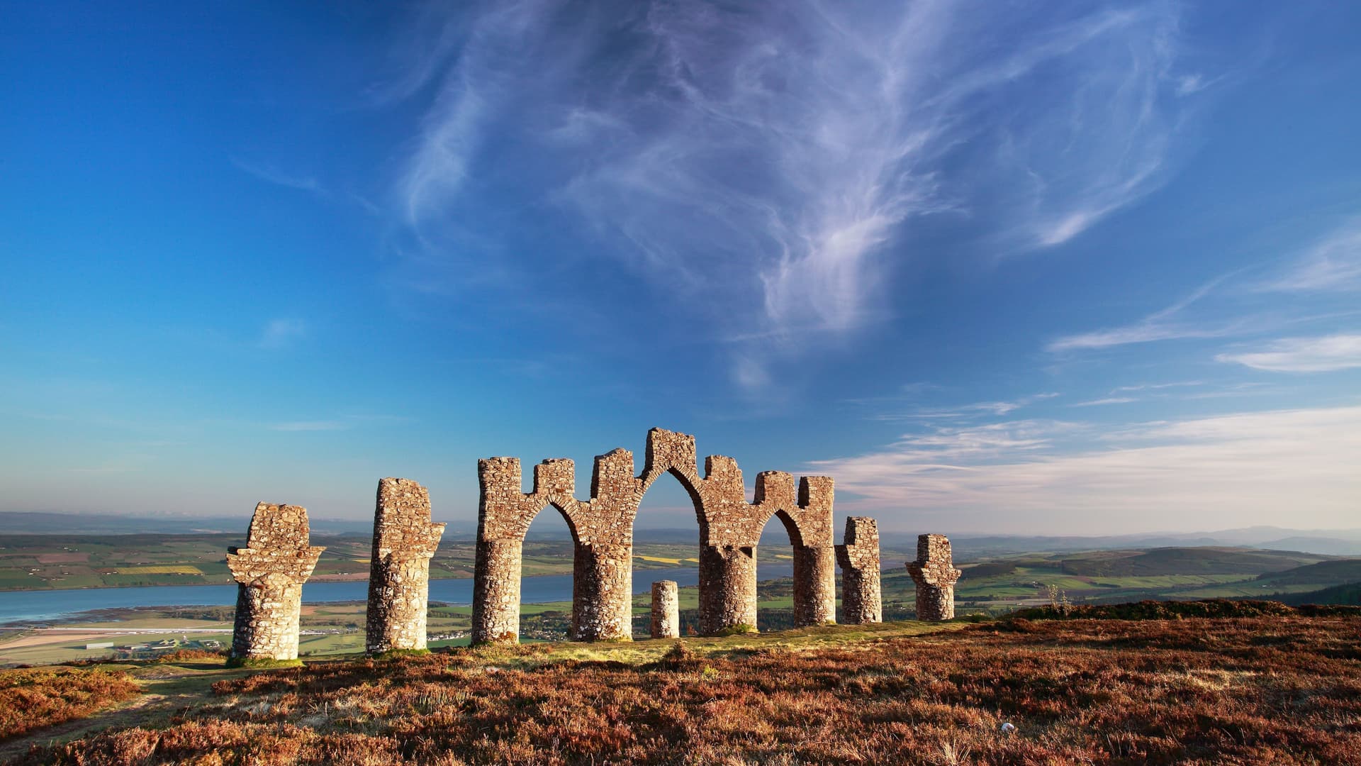 A series of stone archways and pillars, known as the Fyrish Monument, stands on a grassy hill overlooking a body of water and a lush valley in Invergordon, Scotland.