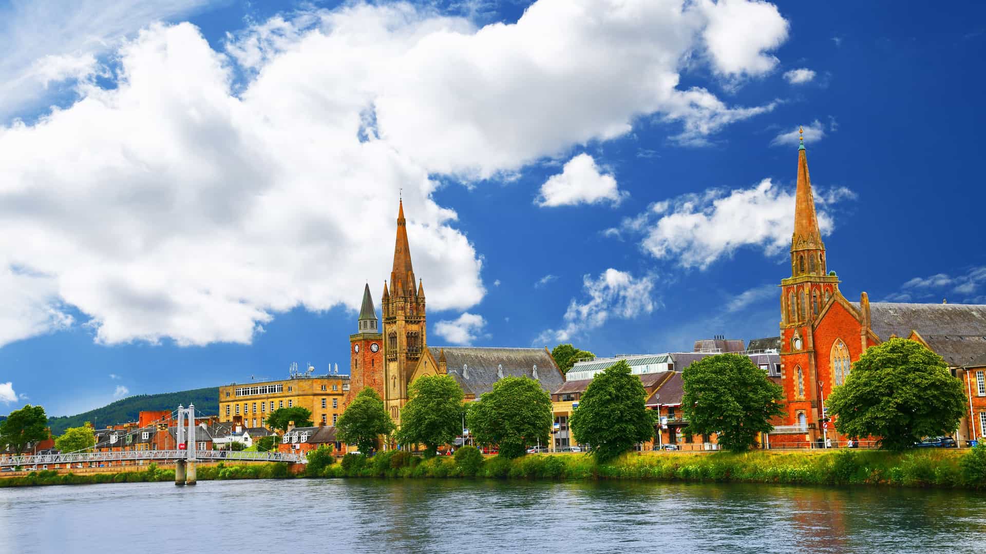 A wide-angle view of the city of Inverness, Scotland, with the River Ness in the foreground and a beautiful skyline featuring several church steeples, a bridge, and buildings on the riverbank.