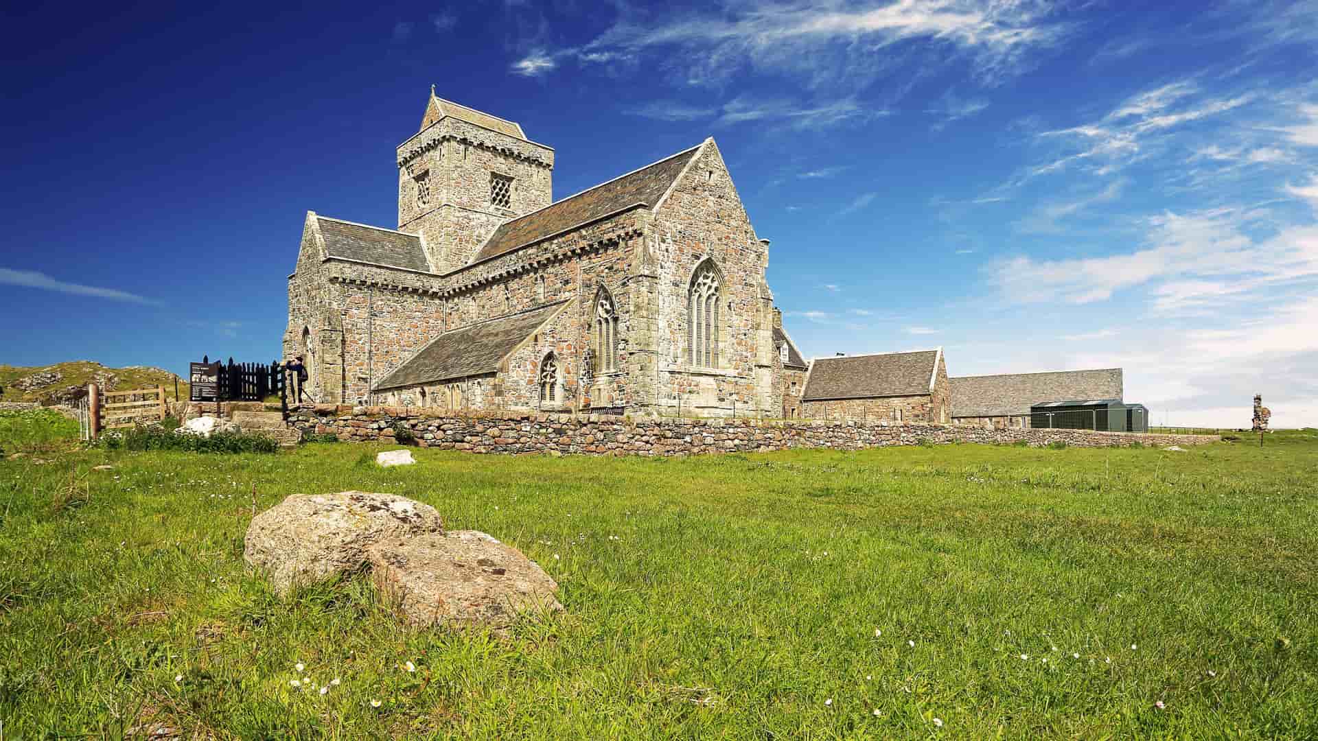 A beautiful shot of the historic Iona Abbey, a large stone church, on the Isle of Iona, Scotland, with green grass in the foreground and a bright blue sky with white clouds overhead.
