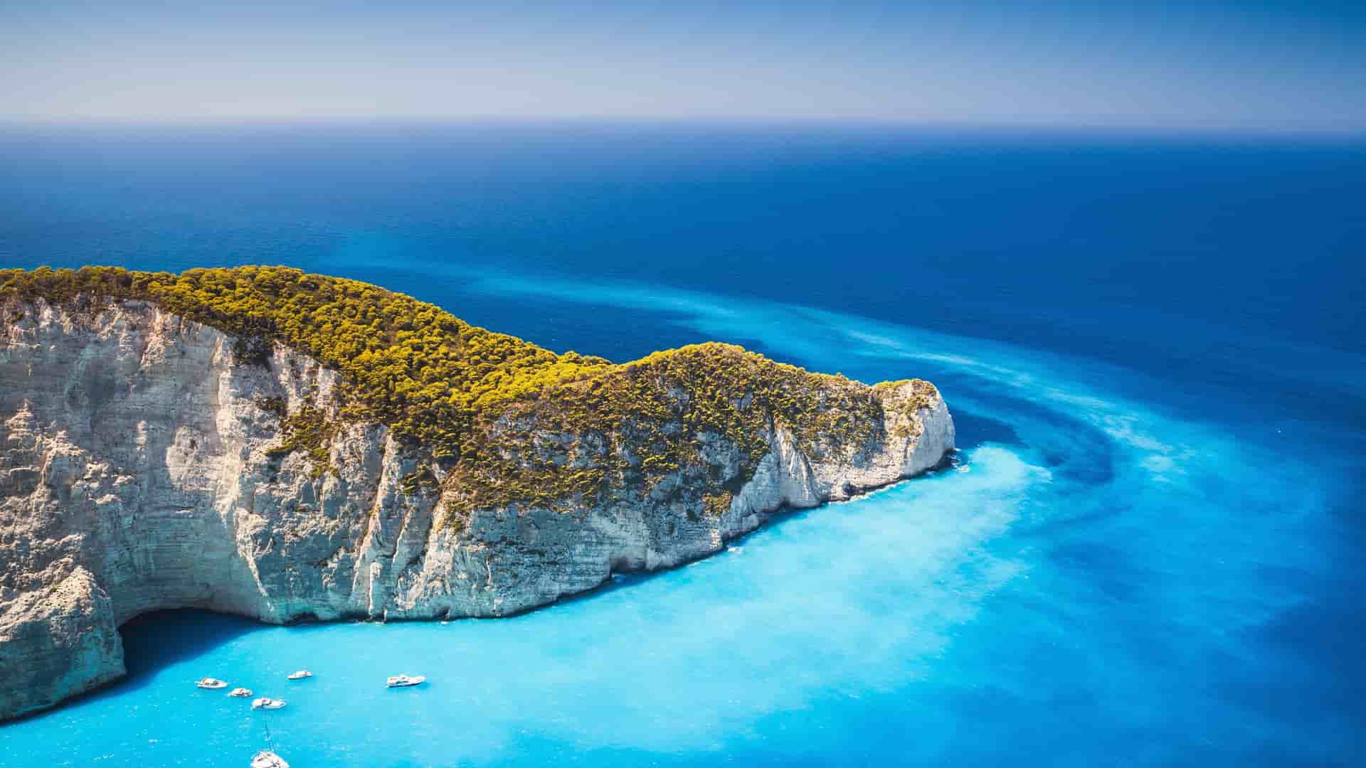 A high-angle drone shot of a lush green cliff face jutting into the brilliant turquoise waters of the Ionian Sea, with small boats anchored below.
