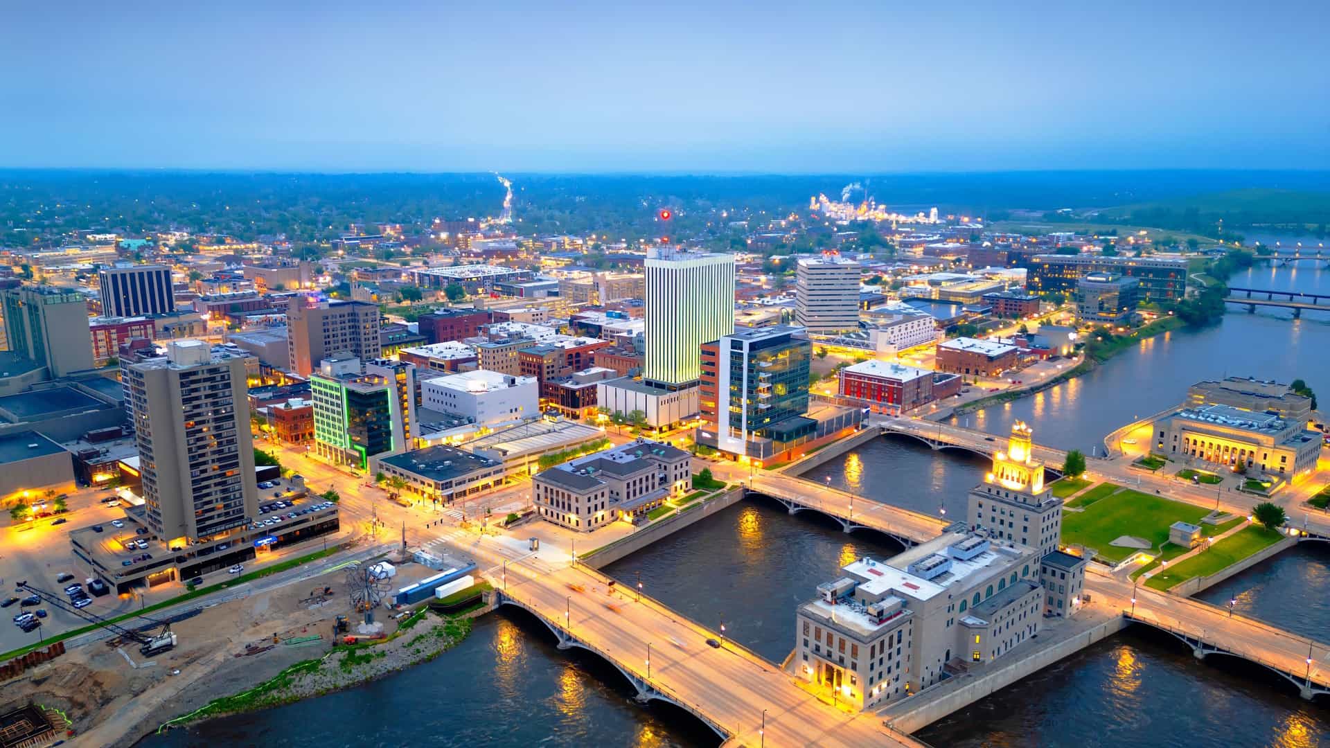 An aerial shot of downtown Cedar Rapids, Iowa, at dusk, with the cityscape illuminated along the river with bridges connecting the two sides.