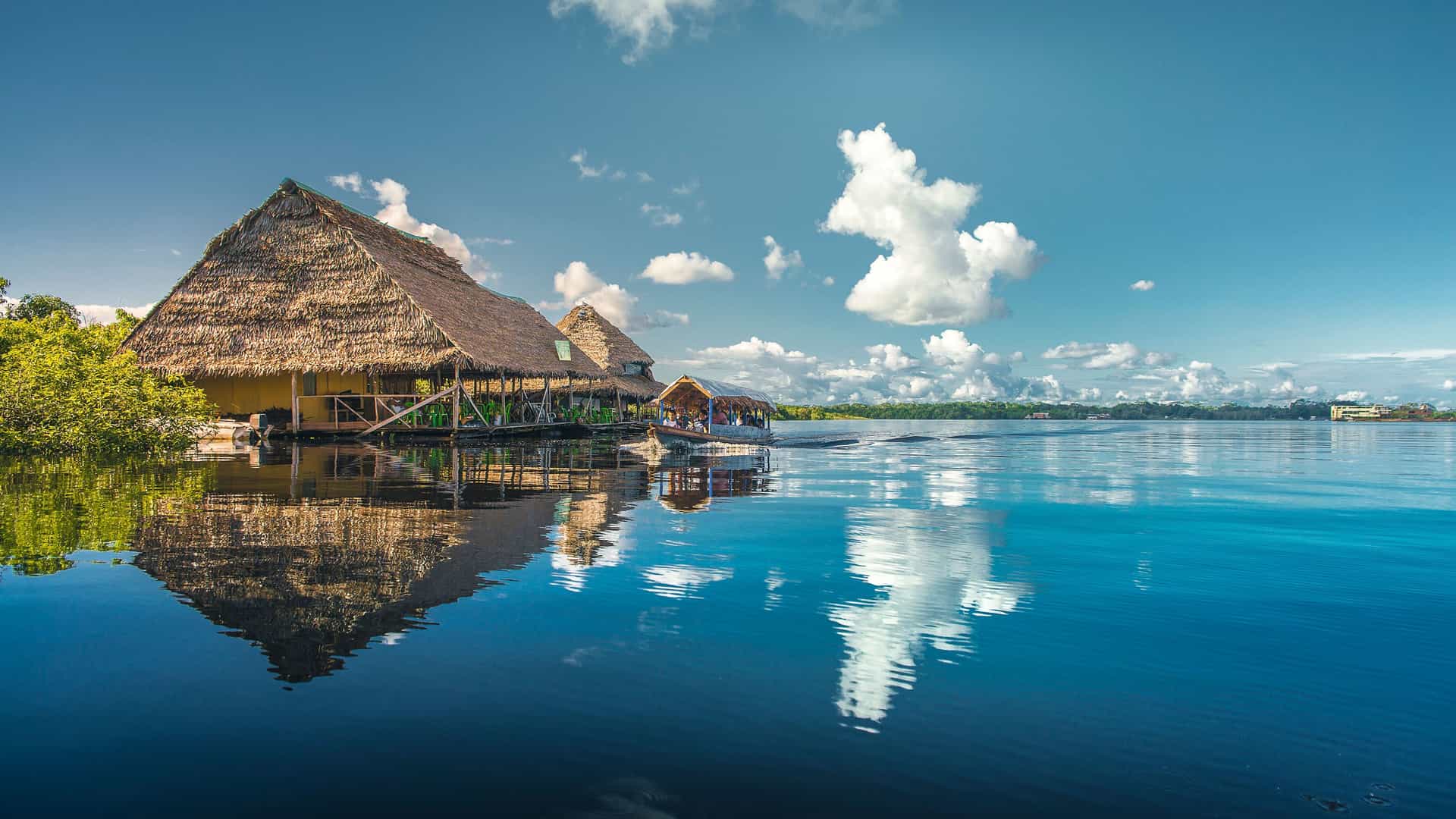A wide-angle landscape shot of a tranquil lake in Iquitos, Peru, with reflections of thatched-roof houses and puffy clouds on the water.