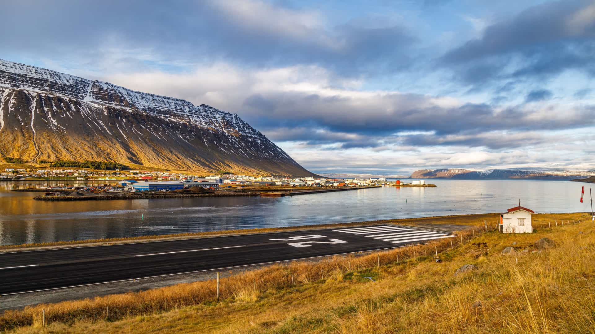 A dramatic coastal scene in Isafjardardjup featuring a runway positioned beside the water, a small building with a red roof and windsock, and a colorful town across the bay backed by a steep snow‑capped mountain.