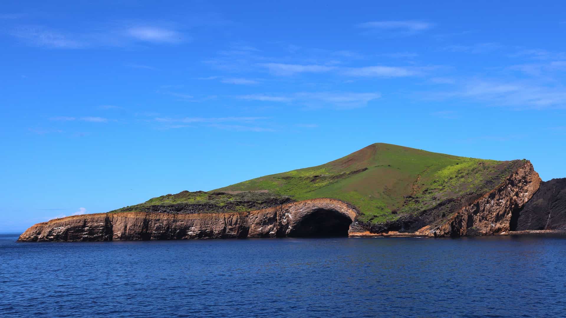 A dramatic view of Vicente Roca Point on Isla Isabela, featuring a large sea cave at the base of the cliffs, with a verdant green hill on top.