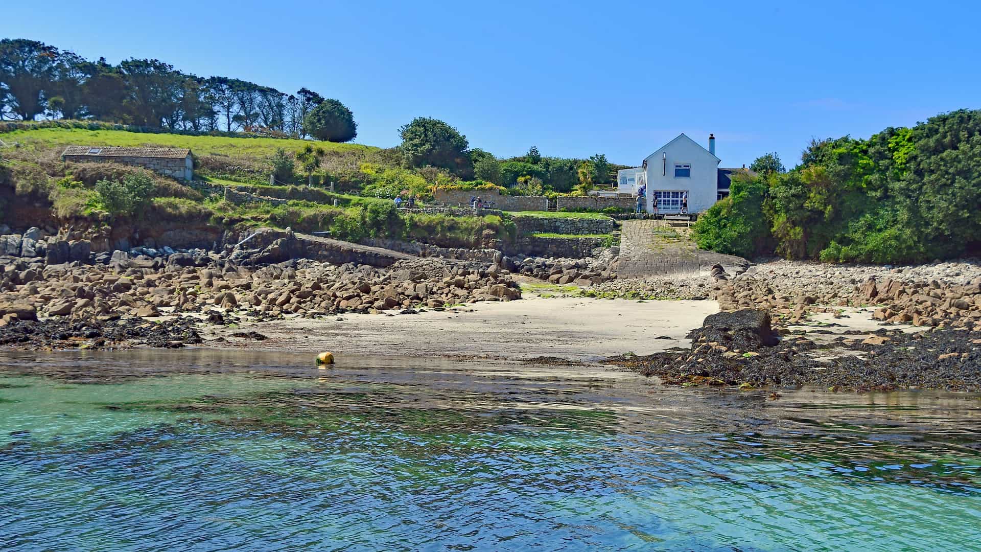 A serene view of a white house with a grassy lawn, situated on a rocky beach with calm turquoise water in the Isle of Scilly.