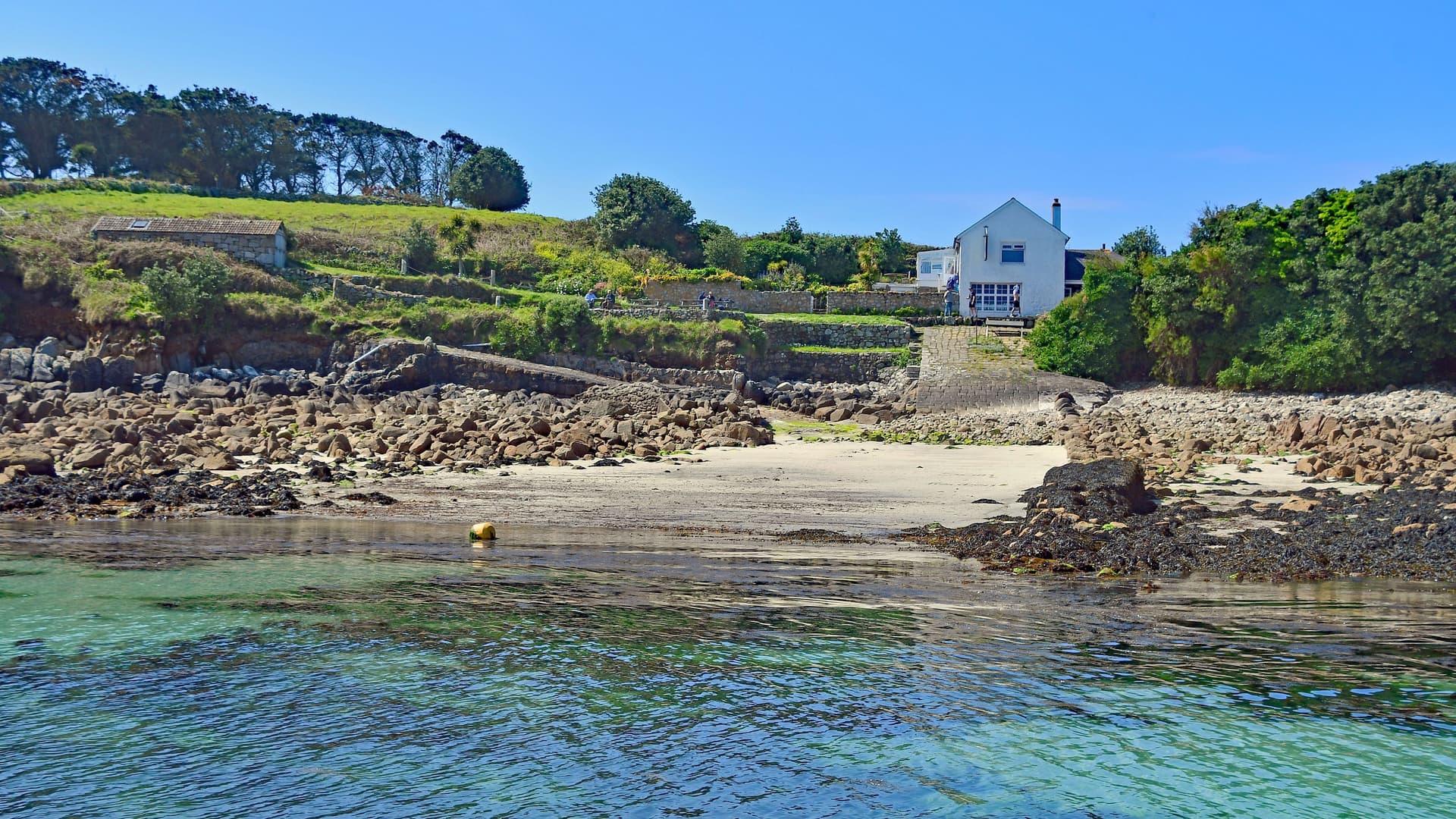 A serene view of a white house with a grassy lawn, situated on a rocky beach with calm turquoise water in the Isle of Scilly.