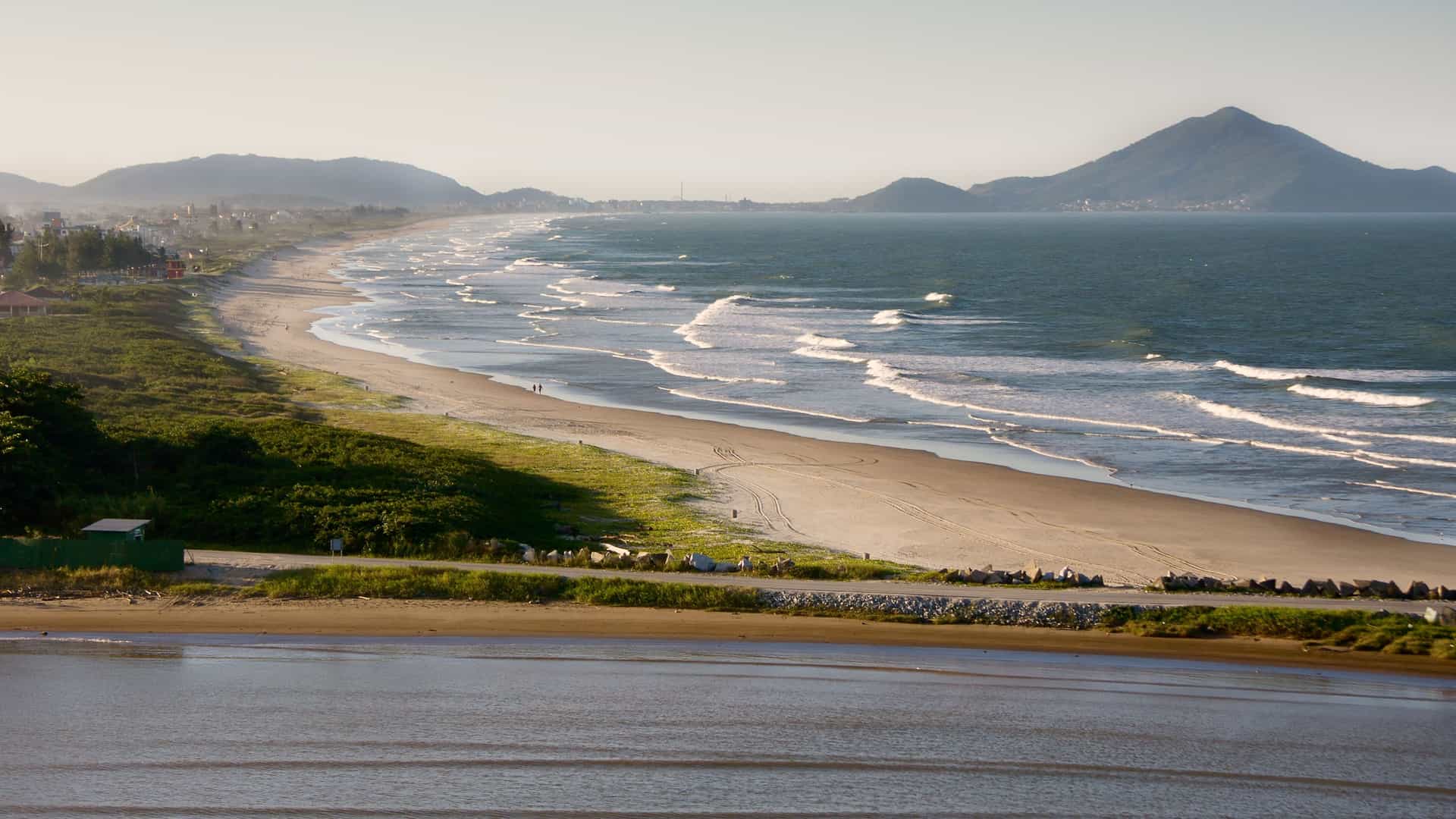 A beautiful golden-hour shot of a long, sandy beach with waves gently rolling onto the shore, with rolling green hills in the background.