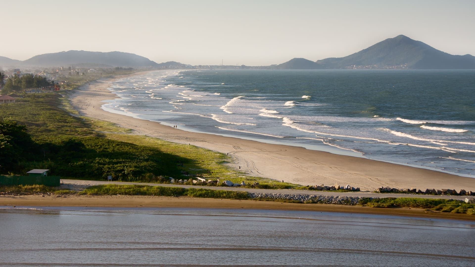A beautiful golden-hour shot of a long, sandy beach with waves gently rolling onto the shore, with rolling green hills in the background.