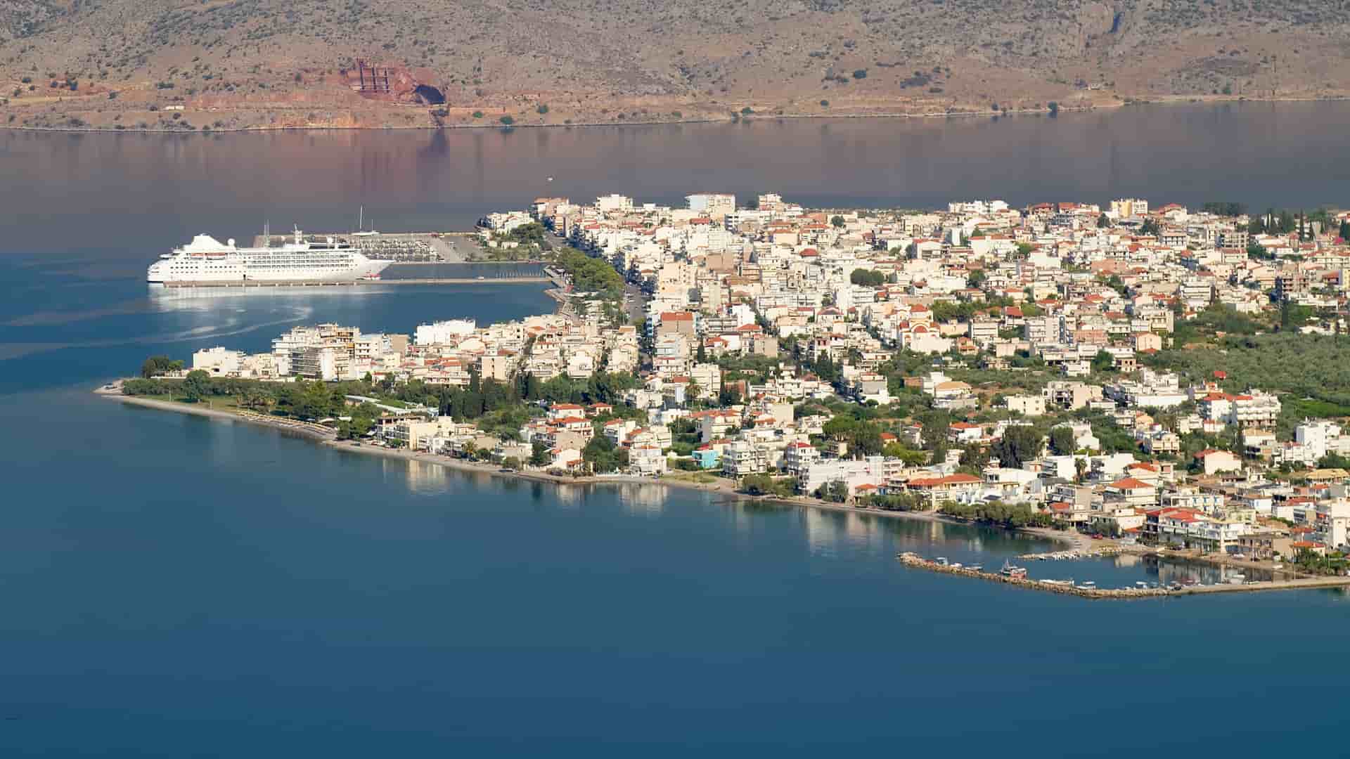 An aerial view of the coastal town of Itea, with a large cruise ship docked at the pier and a dense collection of white buildings stretching along the bay.