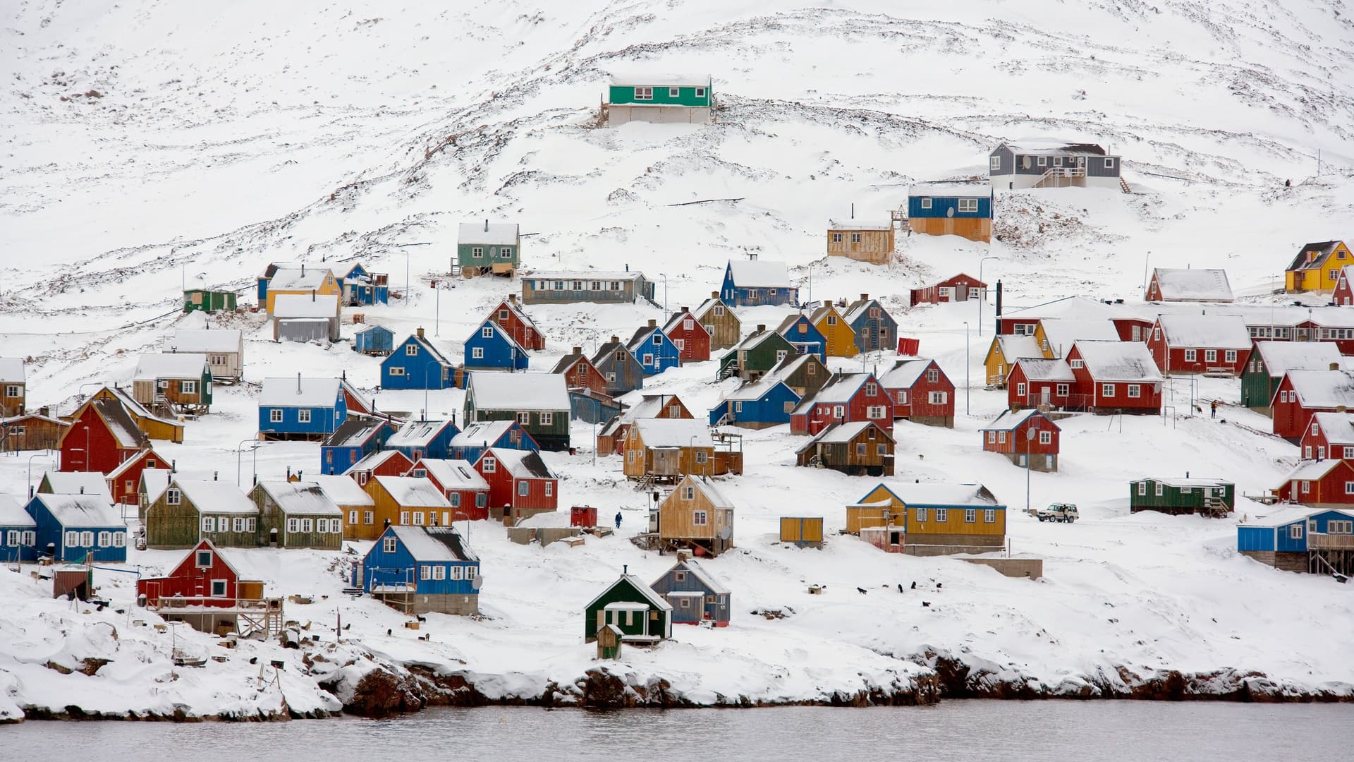 A winter wonderland view of the remote Ittoqqortoormiit village in Greenland, with colorful houses nestled on a snow-covered hillside overlooking the water.