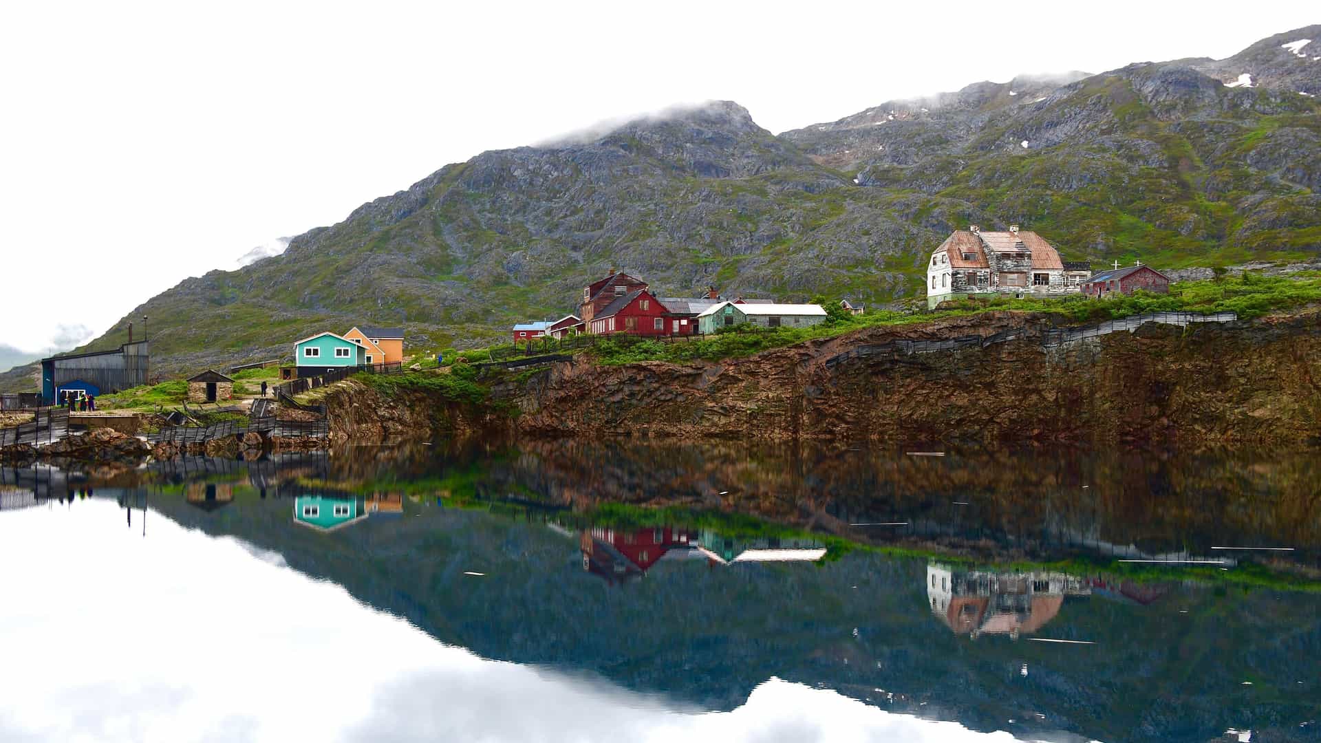 A stunning view of colorful houses perched on a cliffside overlooking a calm, reflective lake, with majestic mountains in the background.