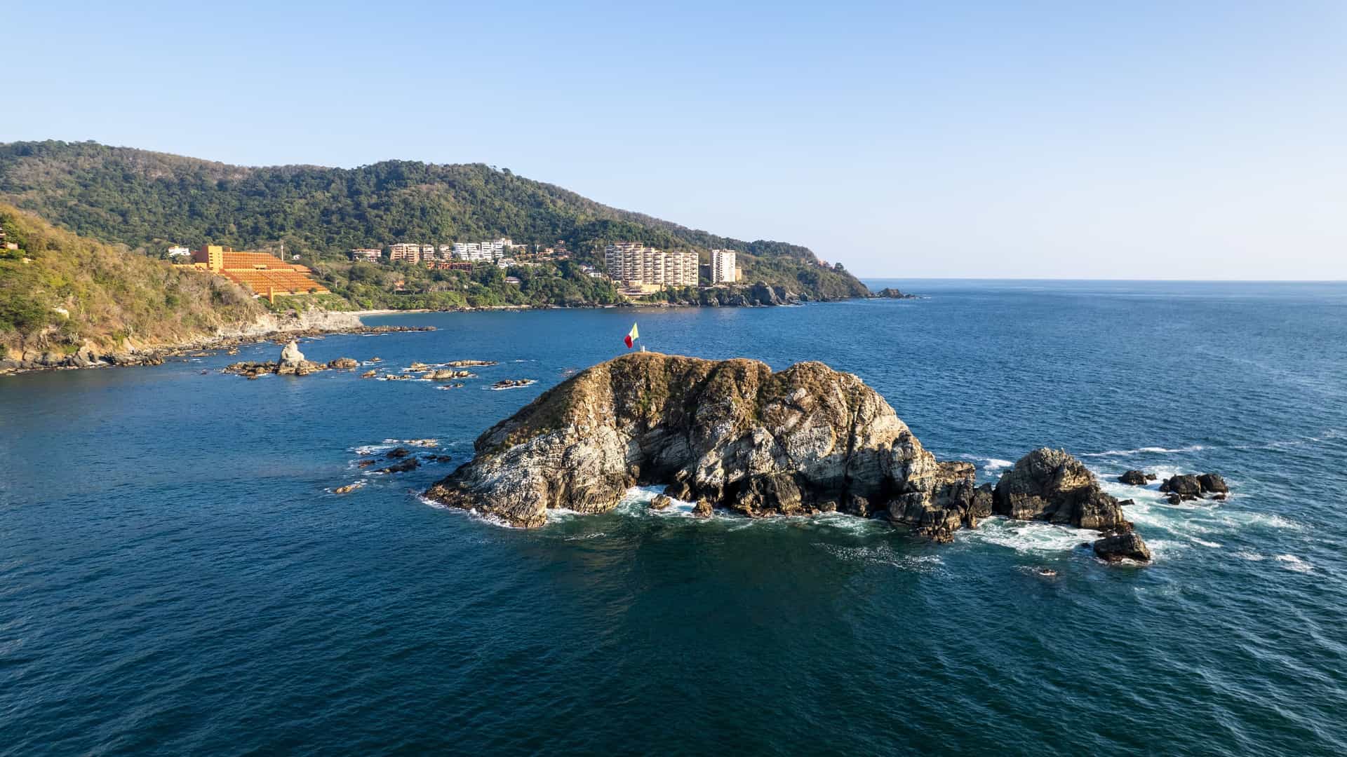 An aerial view of a small rocky island with a Mexican flag on top, off the coast of Ixtapa-Zihuatanejo, with a hillside town and hotels in the background.