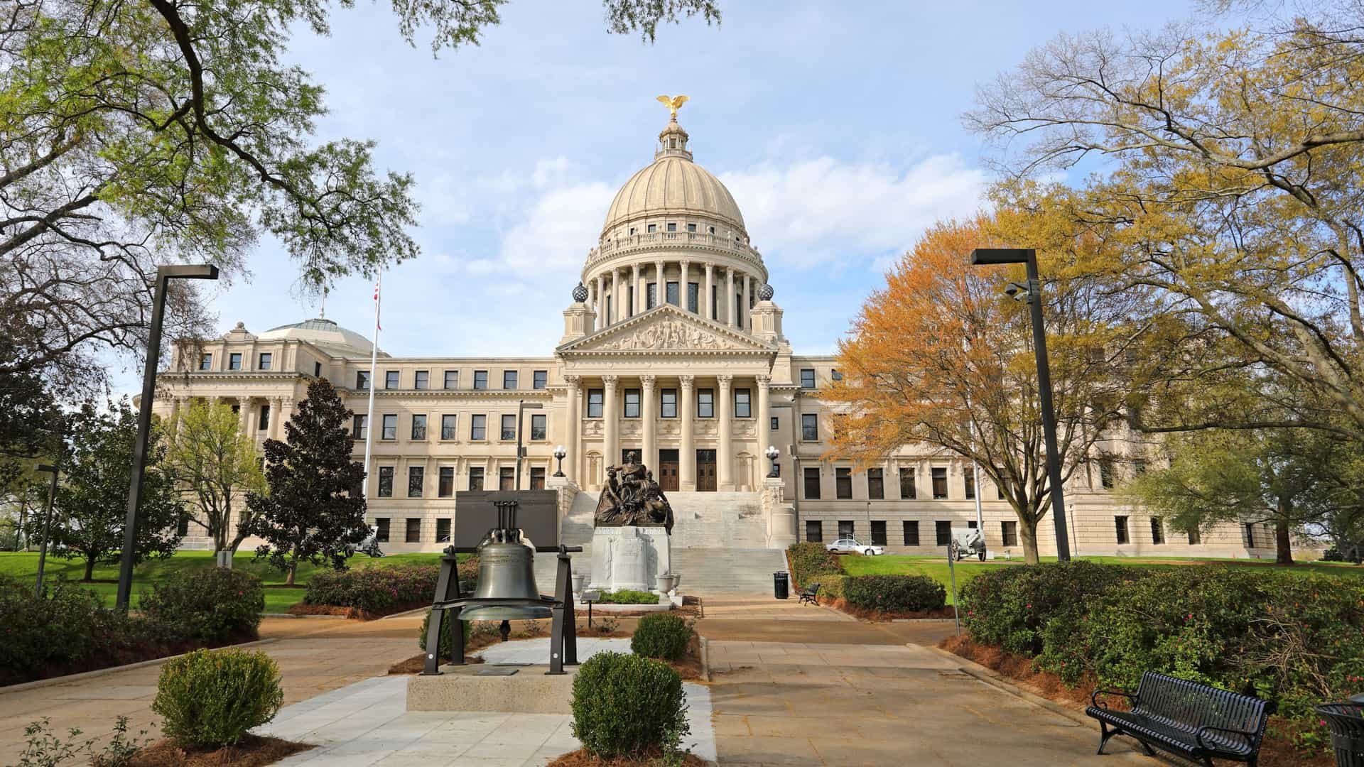 The Mississippi State Capitol Building in Jackson, Mississippi, USA, a grand Beaux-Arts structure with a central dome topped by a golden eagle, viewed from the park pathway that features a historic bell and a bronze statue.