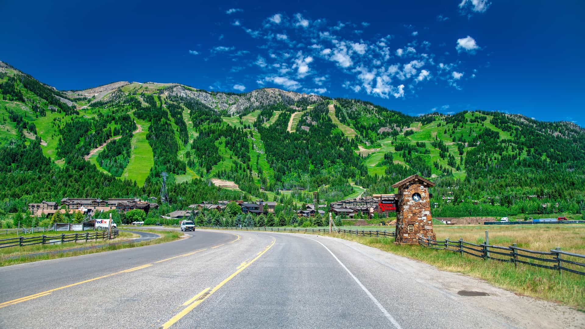 "A picturesque and winding two-lane road in Jackson Hole, Wyoming, leading towards the Teton mountain range with a prominent stone clock tower on the roadside. "