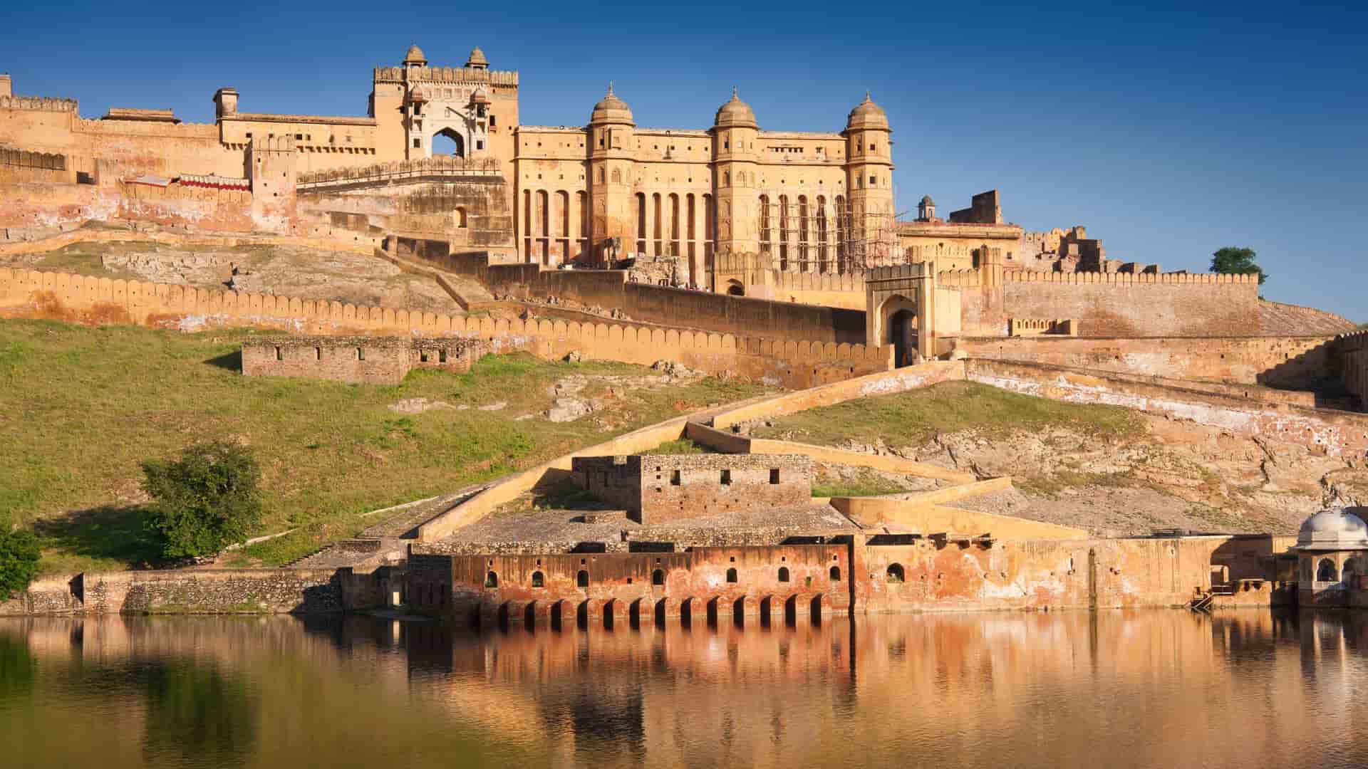 A wide-angle view of the majestic Amber Fort and Palace in Jaipur, India, with its ornate walls and towers reflecting in the serene Maota Lake.