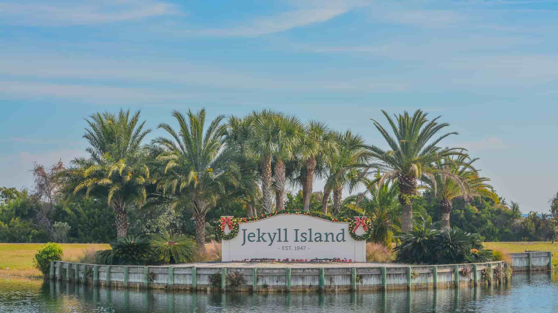 "A welcome sign for Jekyll Island, Georgia, with Christmas decorations on the front, surrounded by lush palm trees and water in the foreground. "