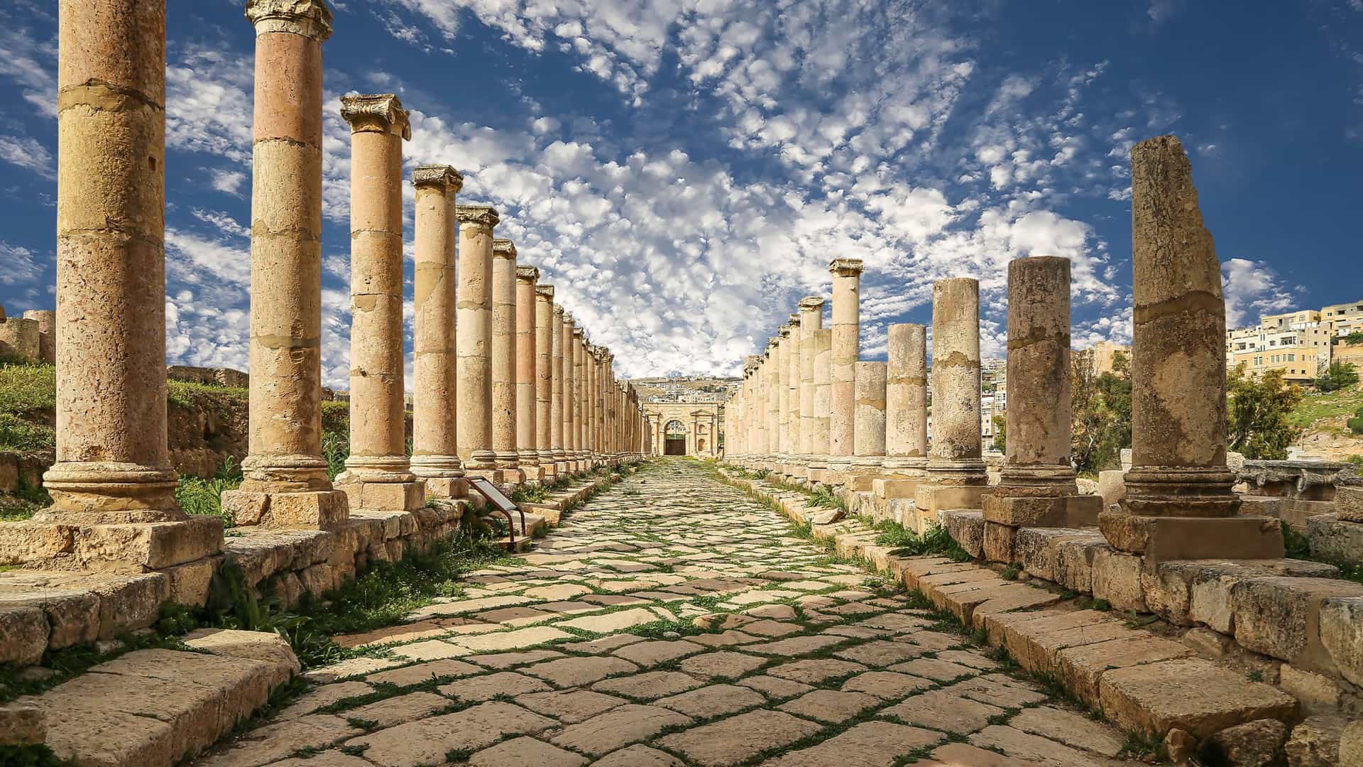 A grand view of the colonnaded street of the ancient Roman ruins of Jerash, with a beautiful blue sky and fluffy clouds overhead.