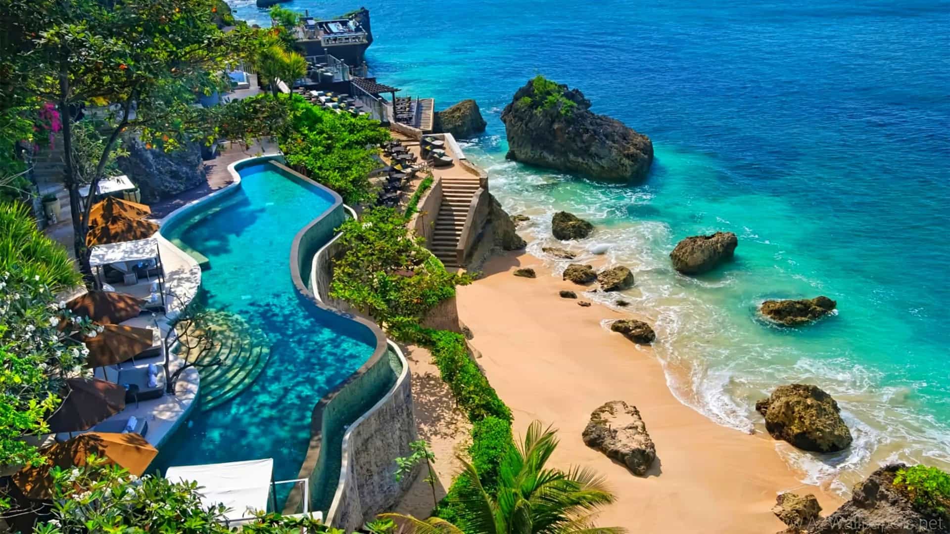 High-angle view of a luxury resort in Jimbaran, Bali, Indonesia, showing a winding, tiered infinity pool built into a cliffside, overlooking a golden-sand beach, rocky outcrops, and the vibrant turquoise waters of the Indian Ocean.