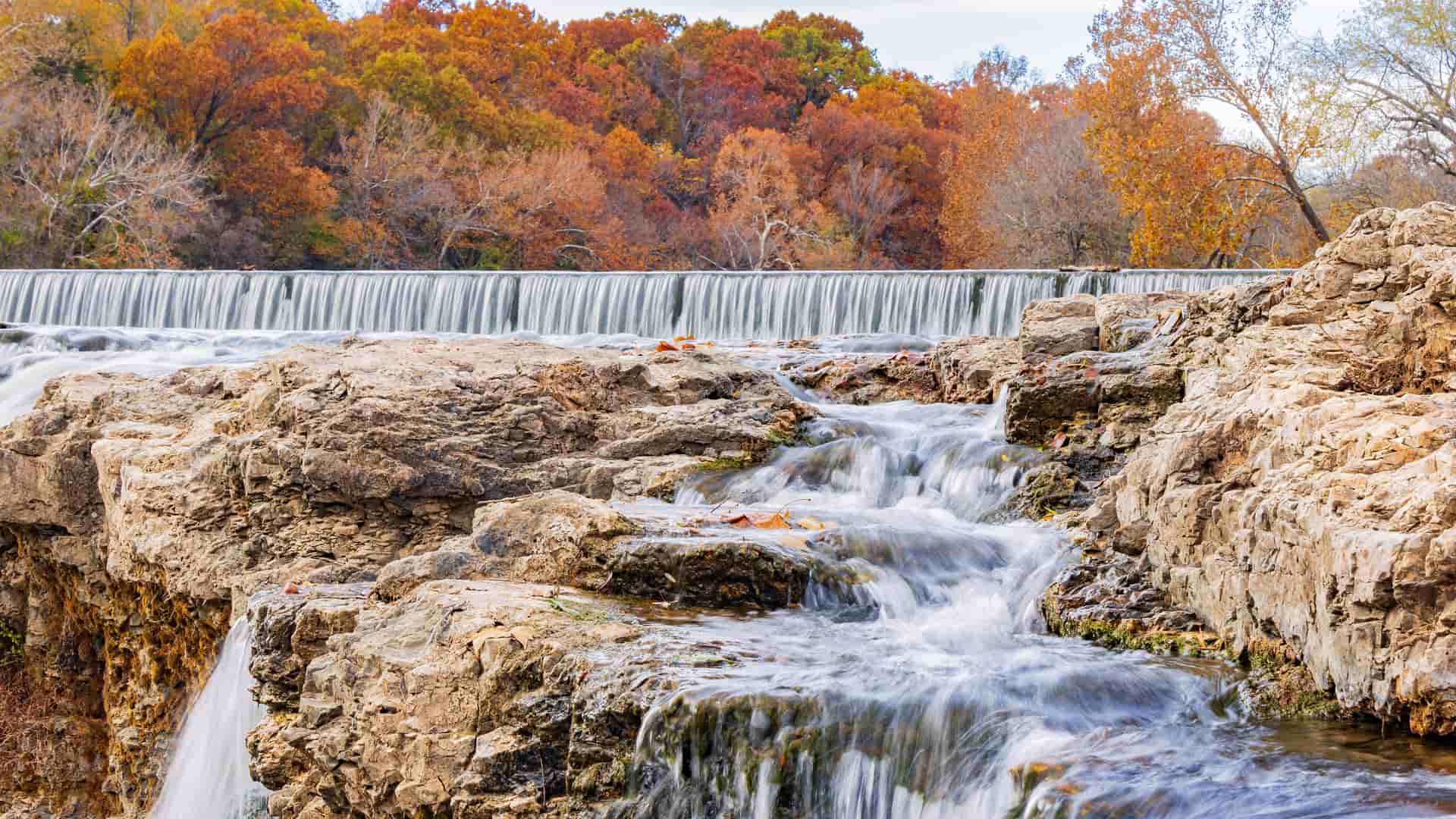 "A stunning autumn landscape shot of the Grand Falls waterfall on Shoal Creek in Joplin, Missouri, with water cascading over the rocky chert limestone formations and vibrant fall foliage in the background. "