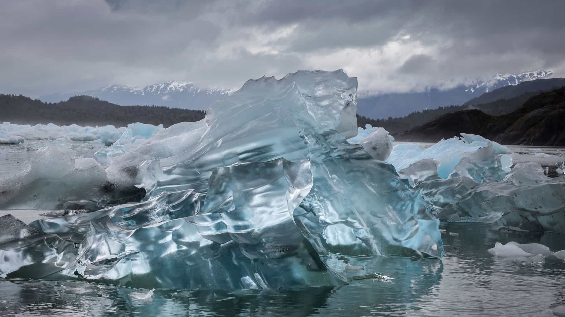 A dramatic close-up of a large, jagged iceberg floating in the water, with a backdrop of snow-capped mountains and a cloudy sky.