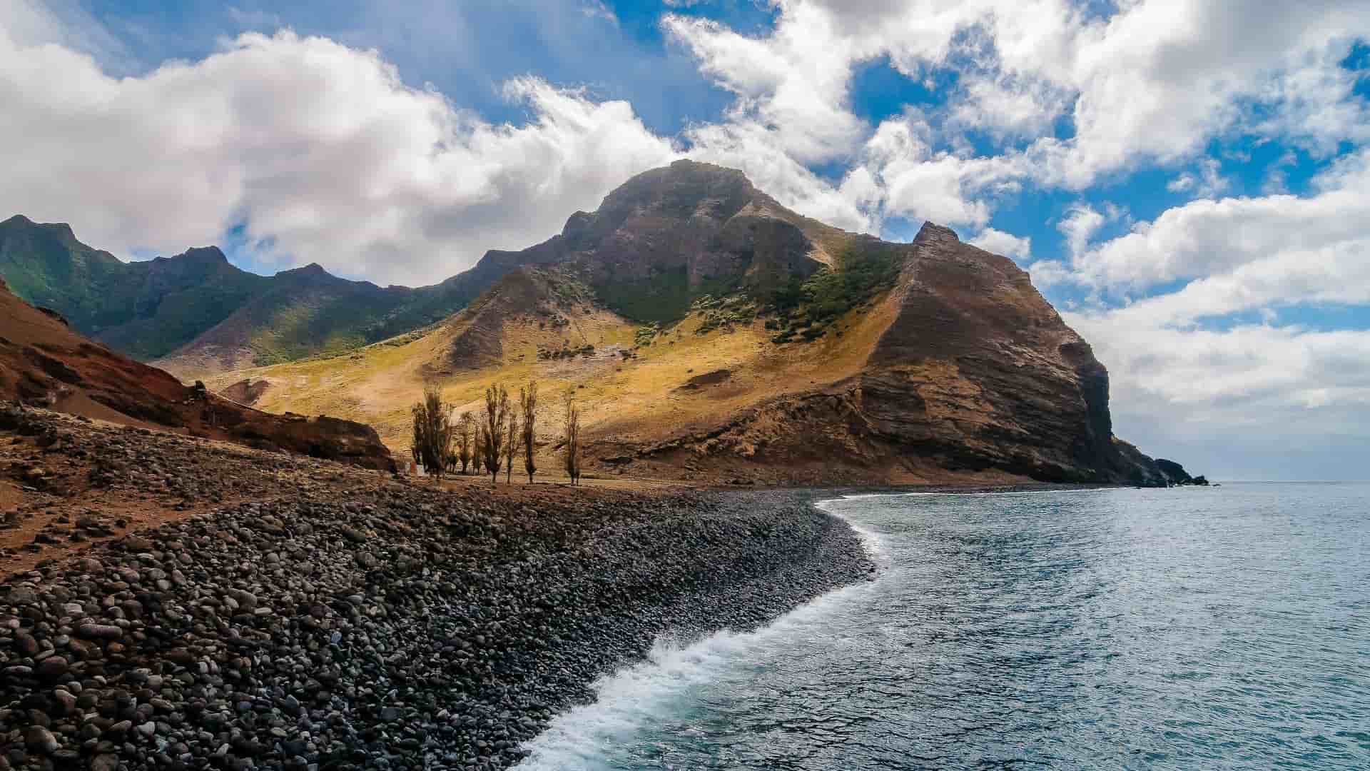 A breathtaking view of Robinson Crusoe Island in the Juan Fernández Islands, featuring a rugged coastline with a black pebble beach, the Pacific Ocean with small waves, and dramatic volcanic mountains under a blue sky with white clouds.