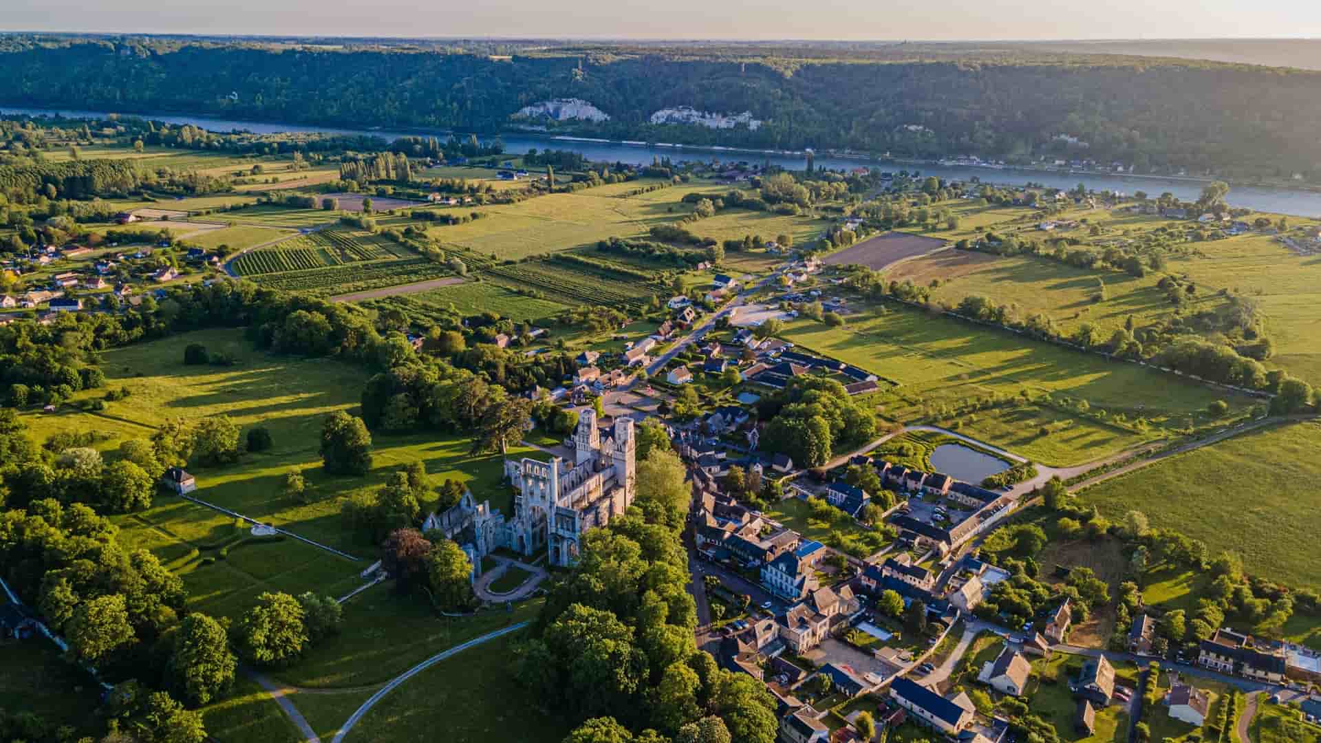 Aerial view of the ruins of Jumièges Abbey surrounded by parkland and the adjacent village of Jumièges, Normandy, France. Farmland extends outward from the village toward the wide, winding Seine River and its chalk cliffs in the distance.