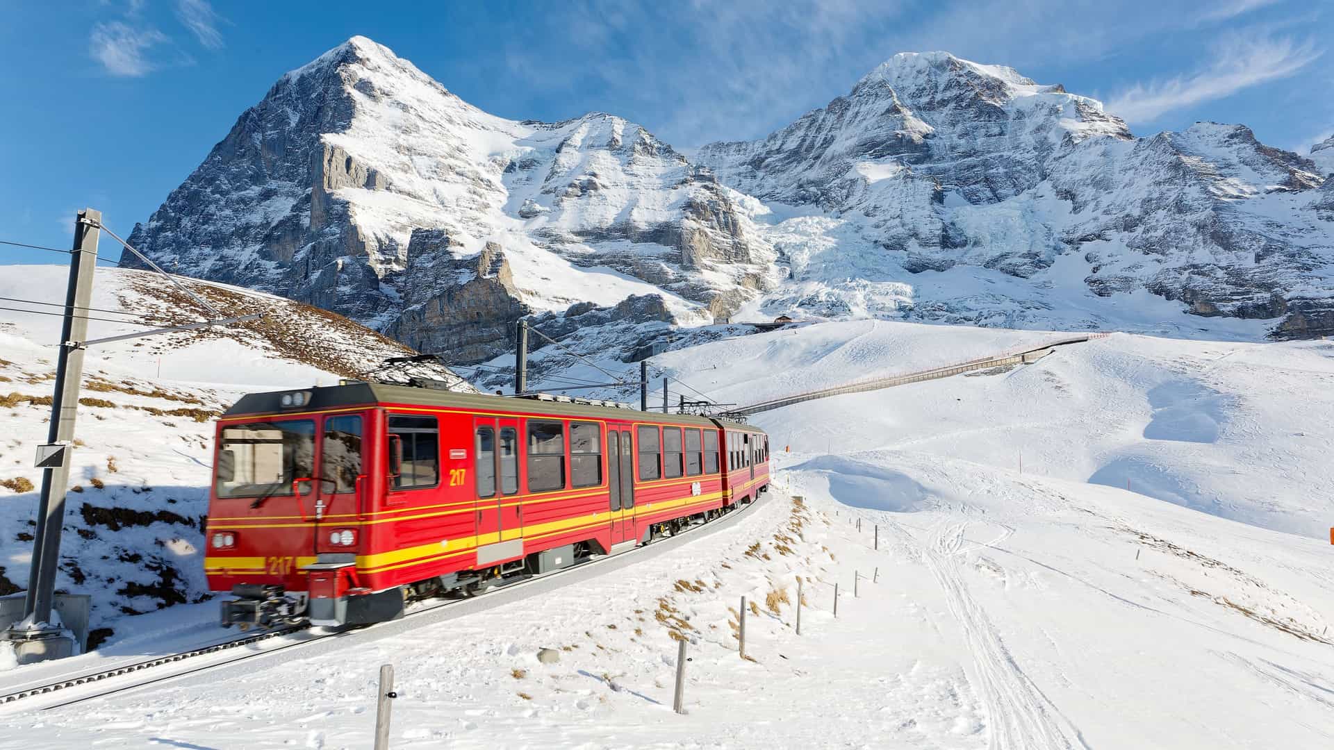A scenic view of a red cogwheel train on the Jungfrau Railway, traveling through snow-covered slopes with the iconic Swiss Alps, Eiger and Mönch mountains, in the background under a clear blue sky.