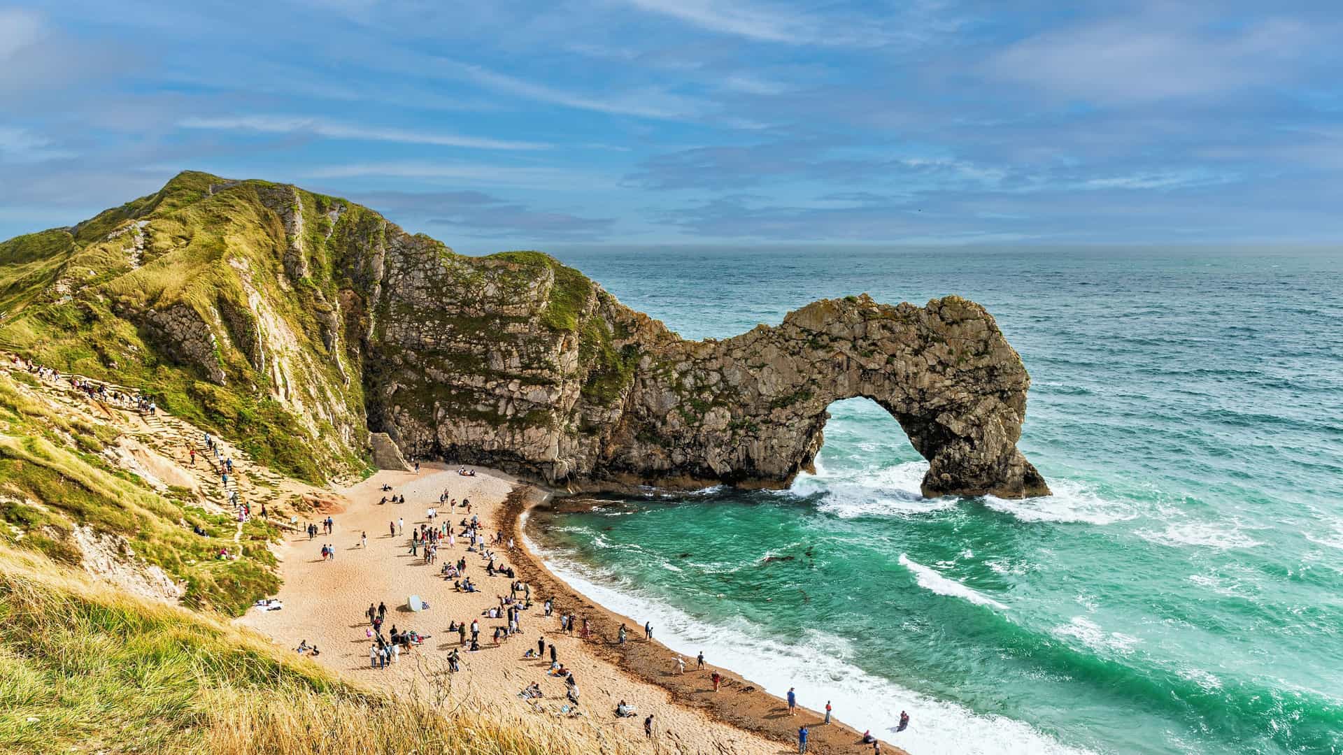 A panoramic view of Durdle Door, a natural limestone arch on the Jurassic Coast, with people enjoying the sandy beach and turquoise water of the English Channel.